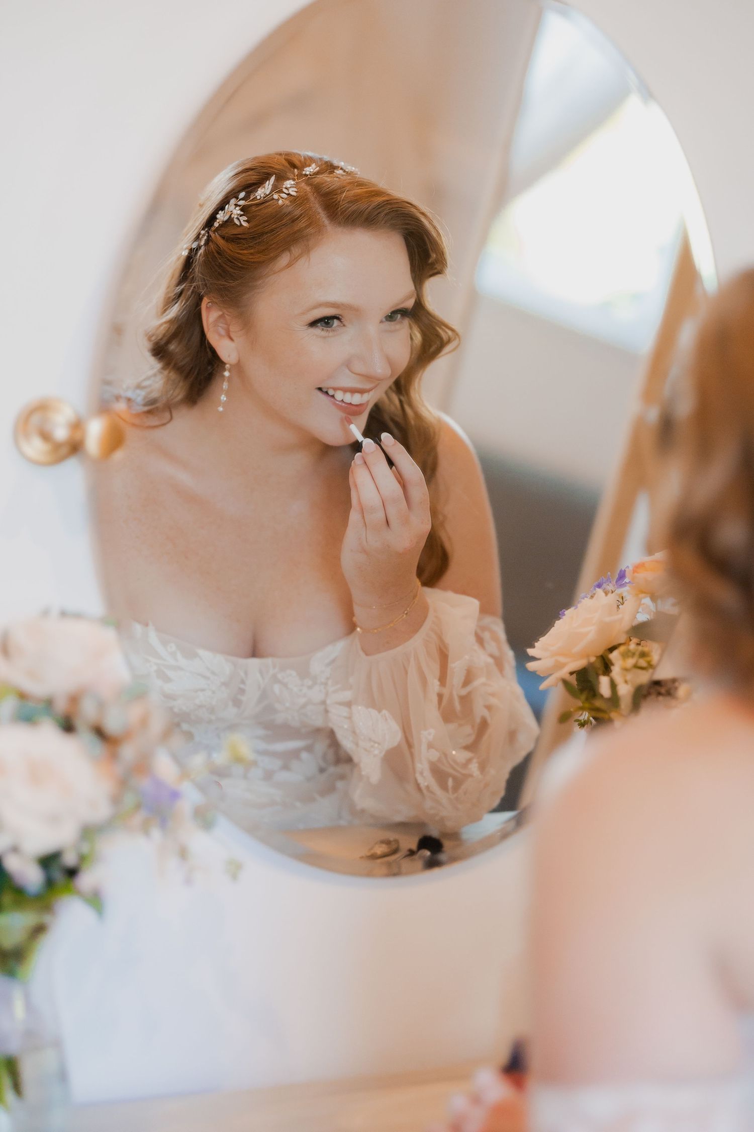 Radiant bride applies lipstick while gazing into ornate mirror on her wedding day.