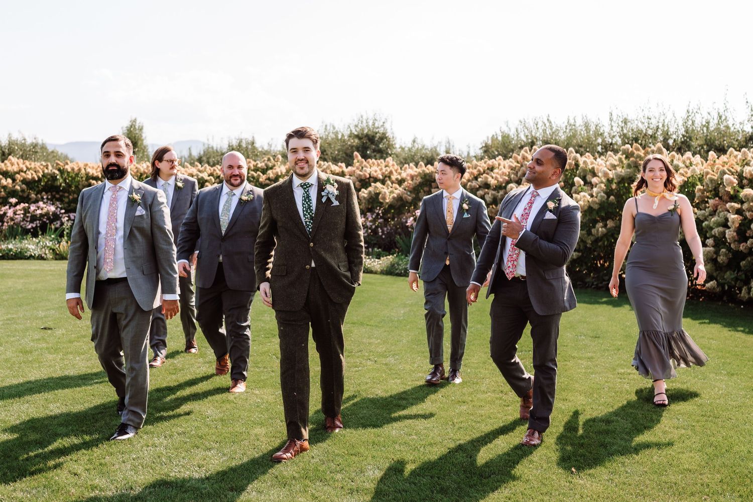 Groom walks confidently with groomsmen across a sunny green lawn at a wedding celebration.