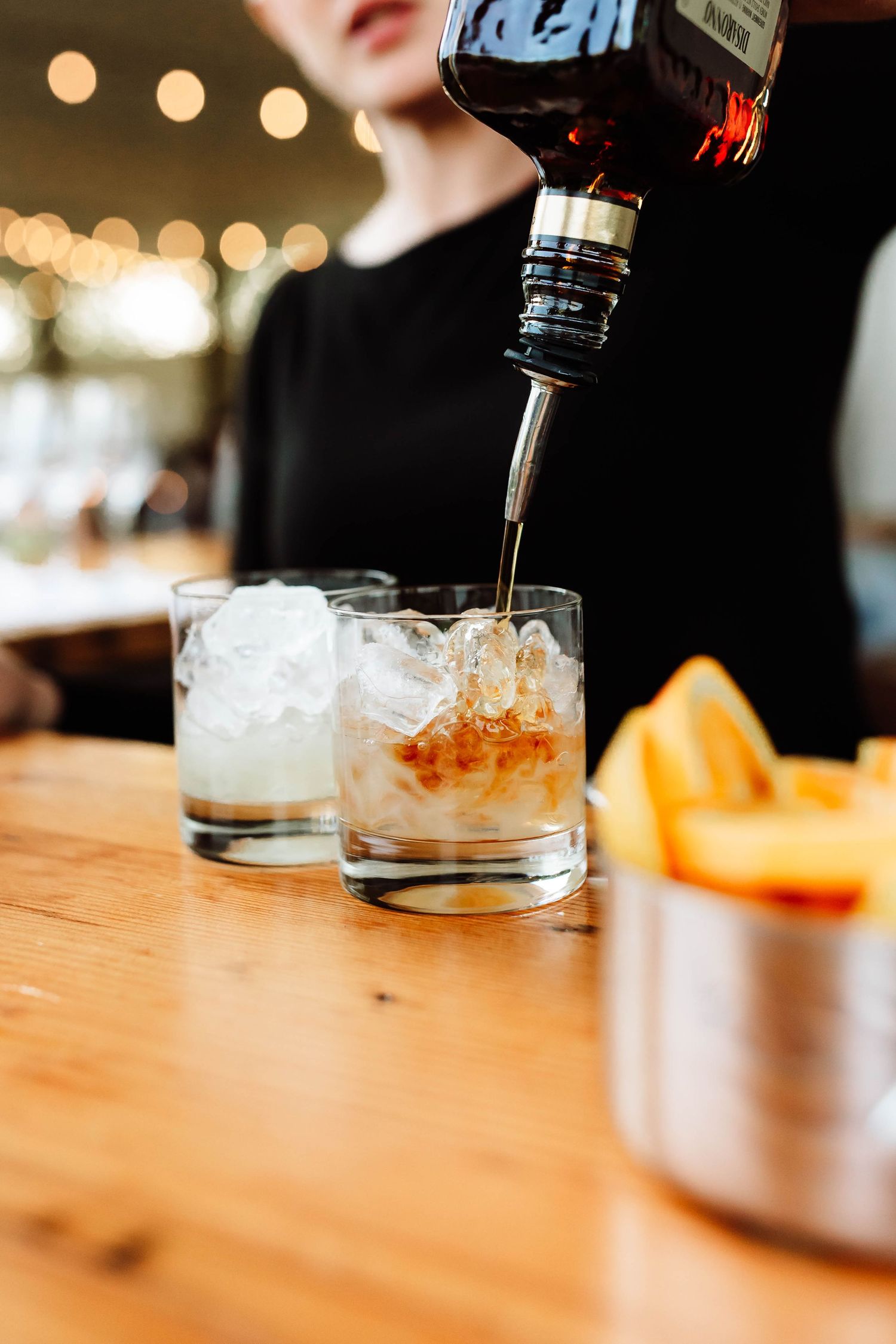 Bartender pouring whiskey over ice into a rocks glass at a wedding bar with citrus garnishes nearby.