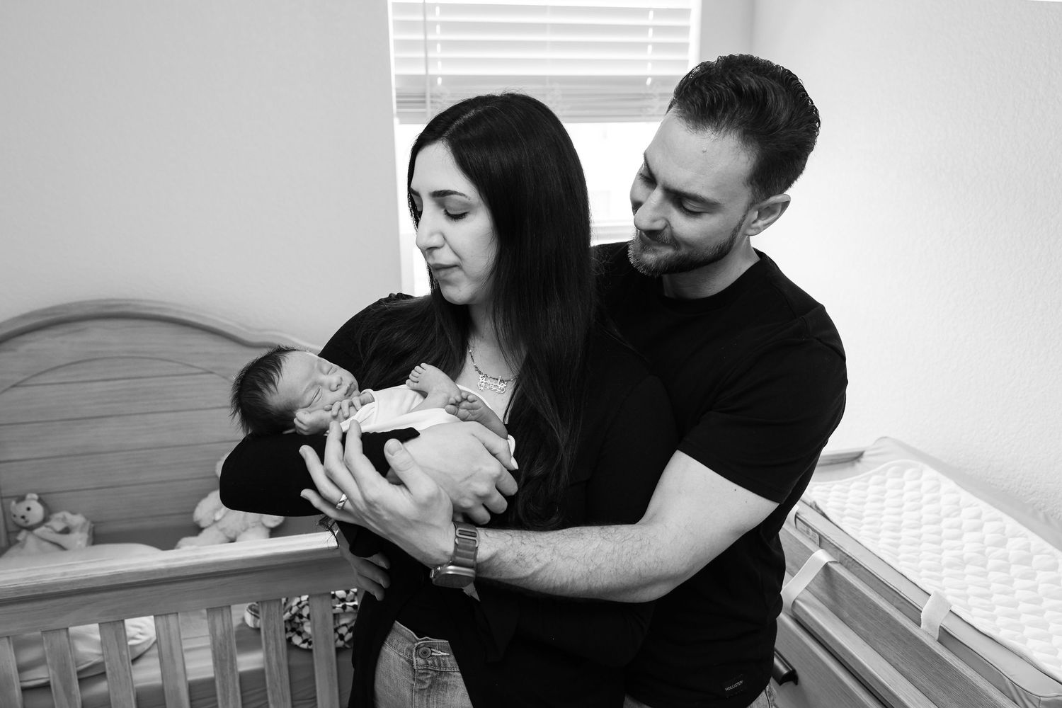 Black and white photo of couple holding newborn baby near crib in nursery.