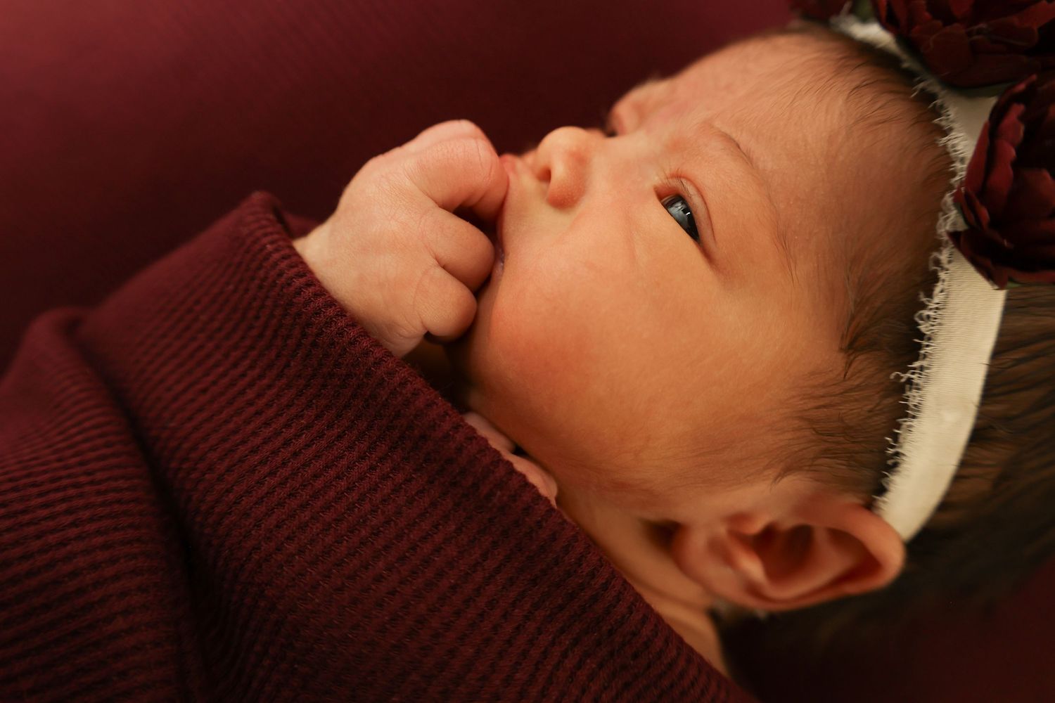 Close-up of a newborn baby with fingers near mouth wearing a maroon outfit and white headband.