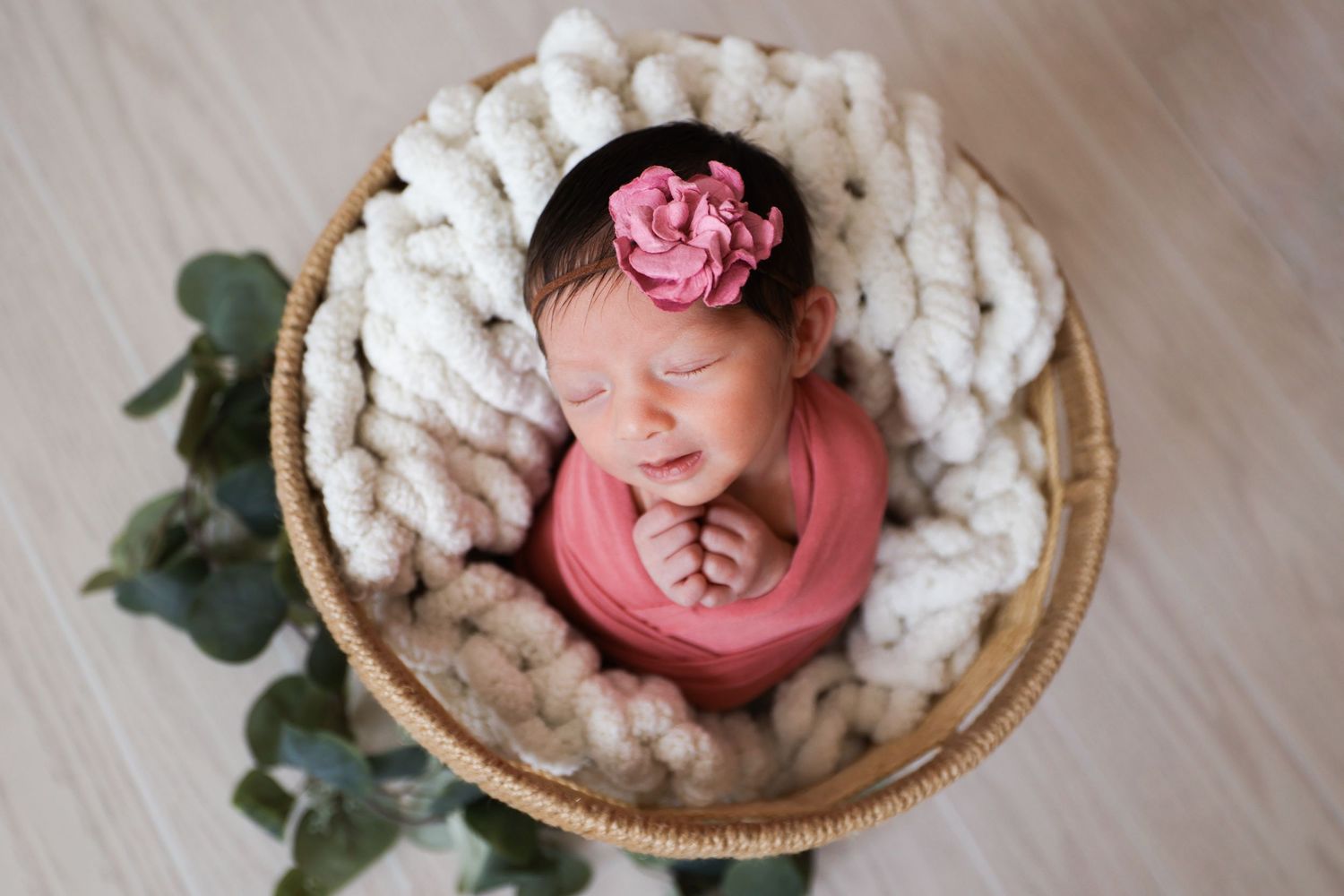Sleeping newborn baby girl in coral wrap and floral headband nestled in a wicker basket.