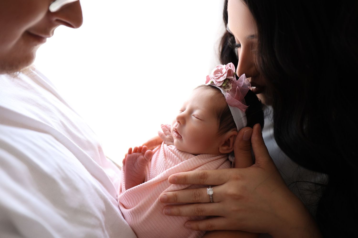 Two women tenderly kissing a sleeping newborn baby girl wrapped in pink with a floral headband.