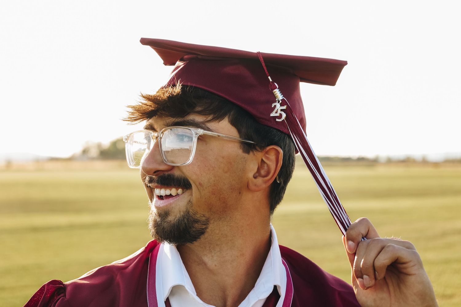 Happy young man in maroon graduation cap and gown laughing outdoors in a field.