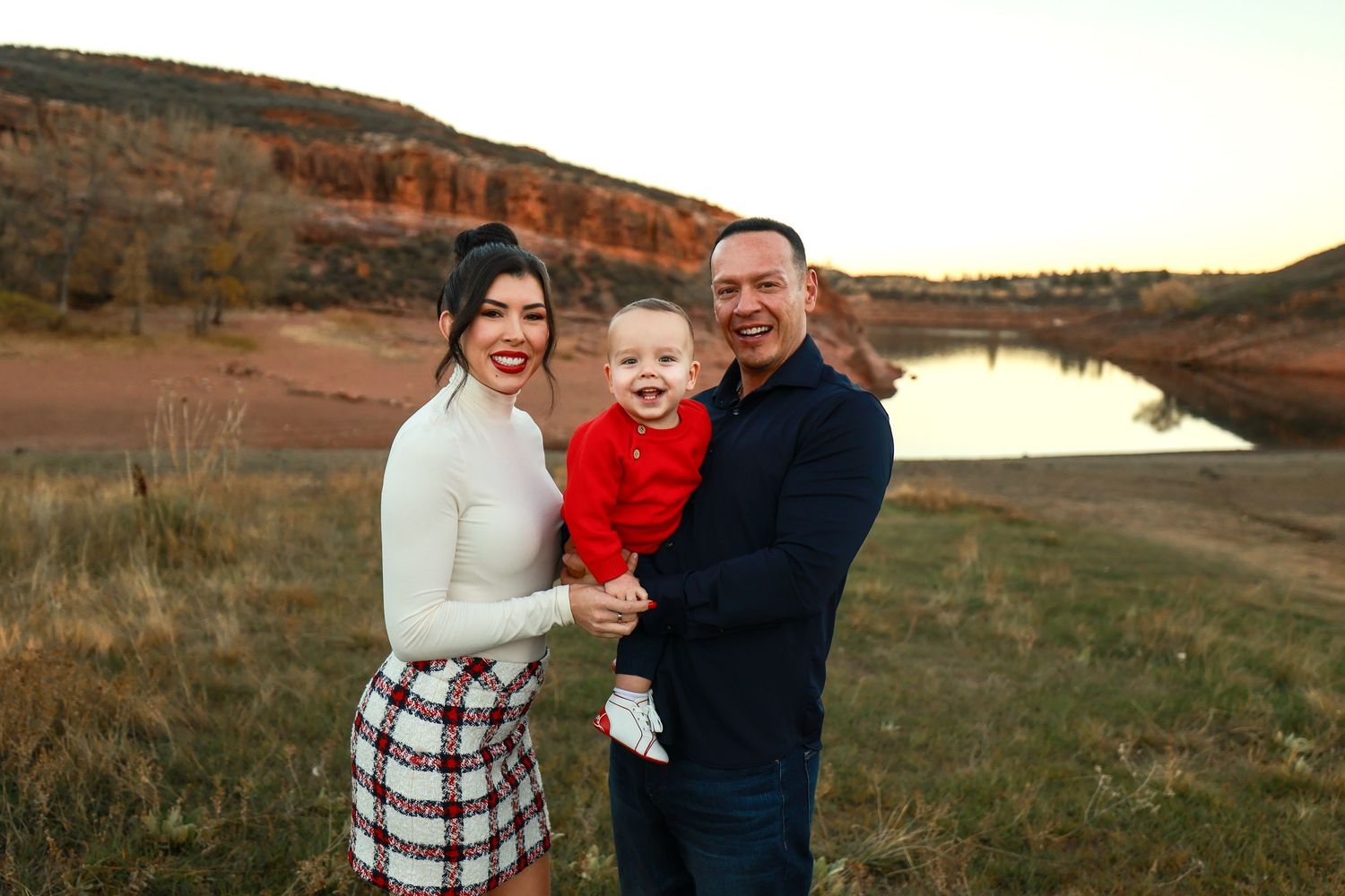 Happy family of three posing outdoors near a red rock lake at sunset with smiling baby in red.
