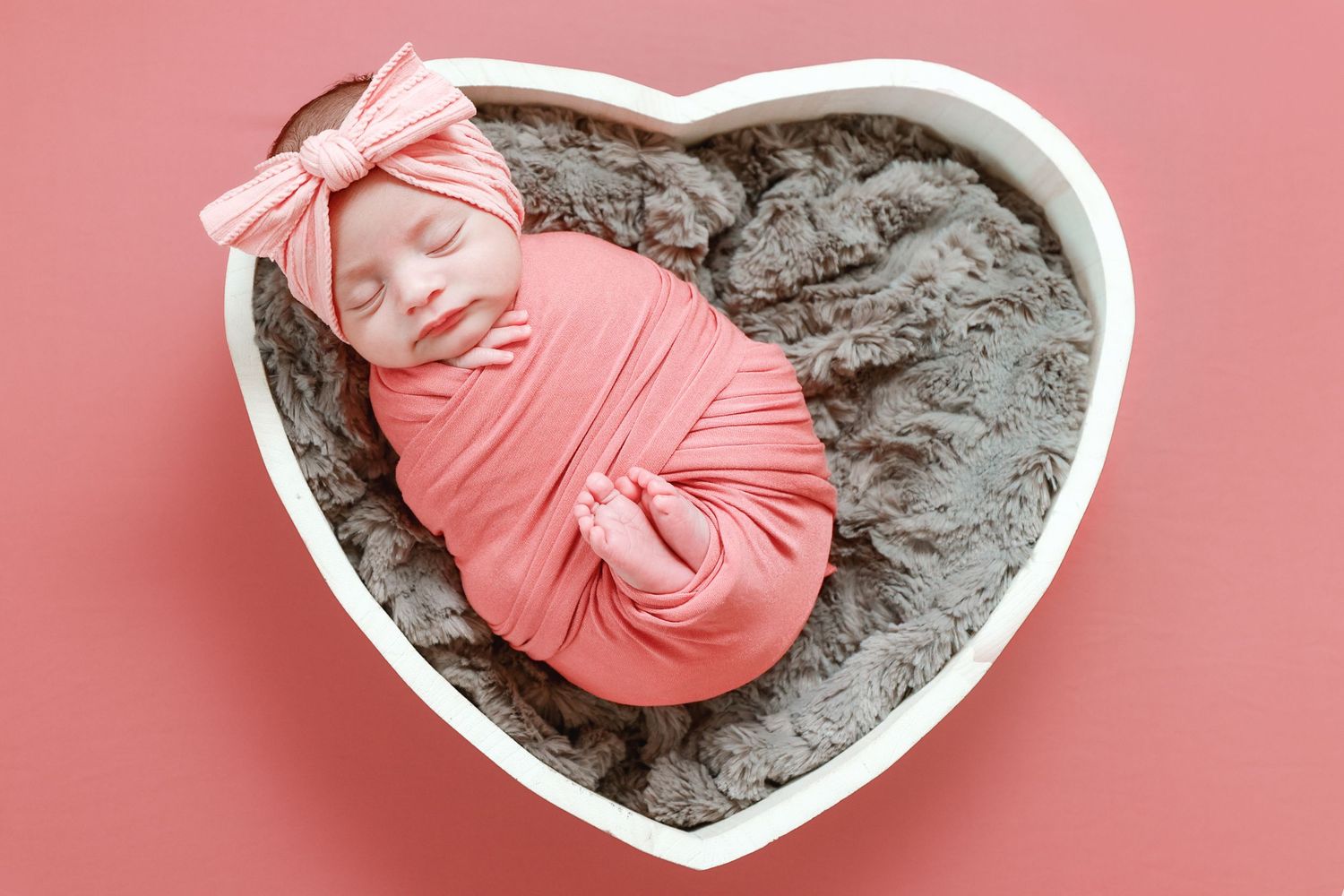 Sleeping newborn baby girl swaddled in coral pink wrap inside white heart-shaped bowl on pink background.