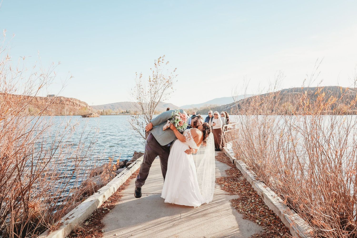Bride and groom kissing on a dock with a scenic lake and mountains behind them.
