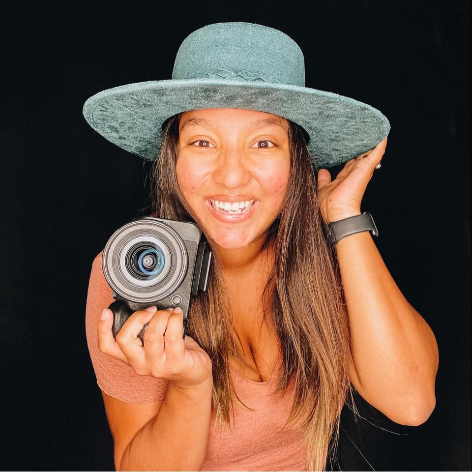 Smiling woman holding a camera wearing a wide-brim hat, professional photographer portrait.