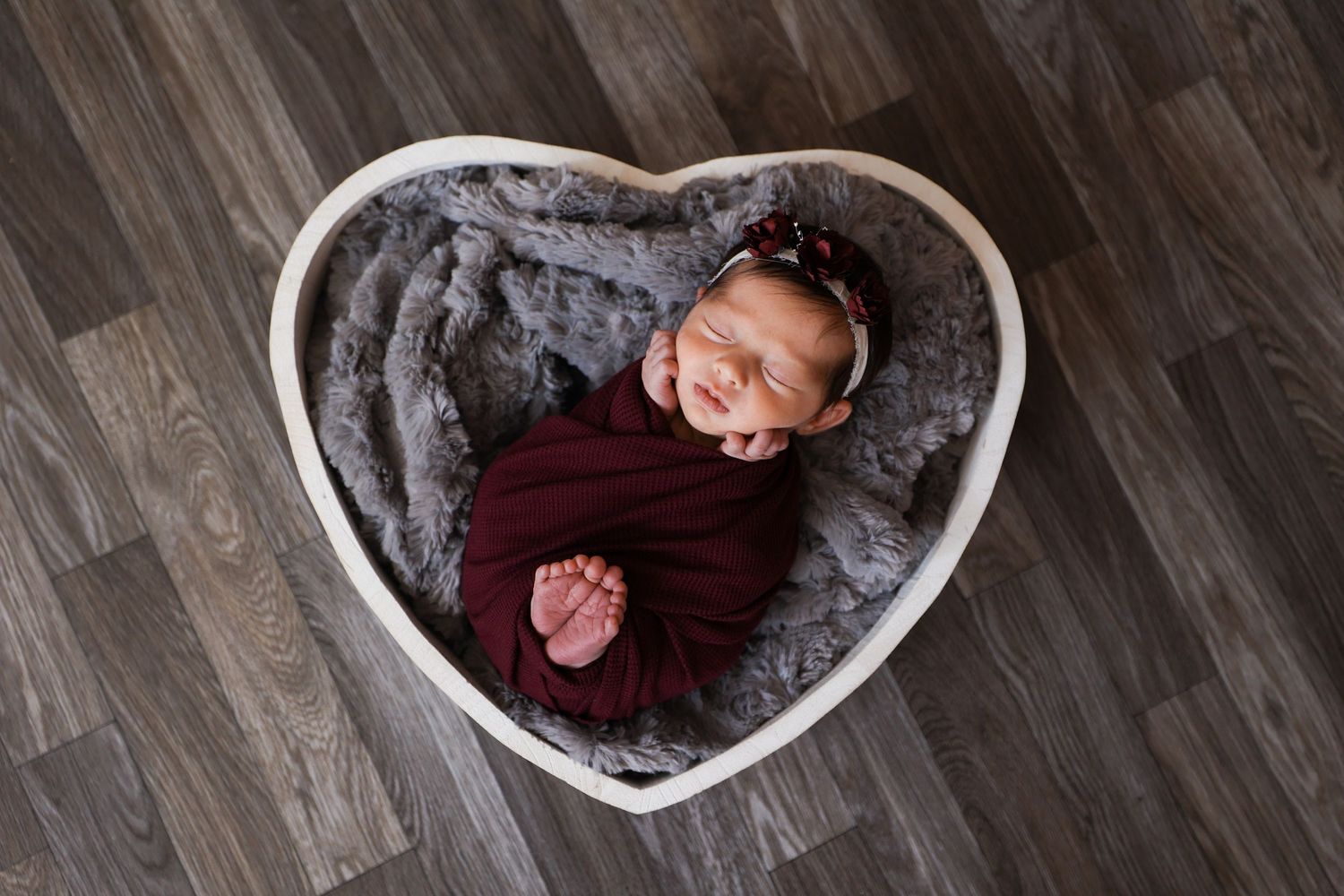 Smiling newborn baby wrapped in burgundy swaddle inside a heart-shaped wooden bowl.