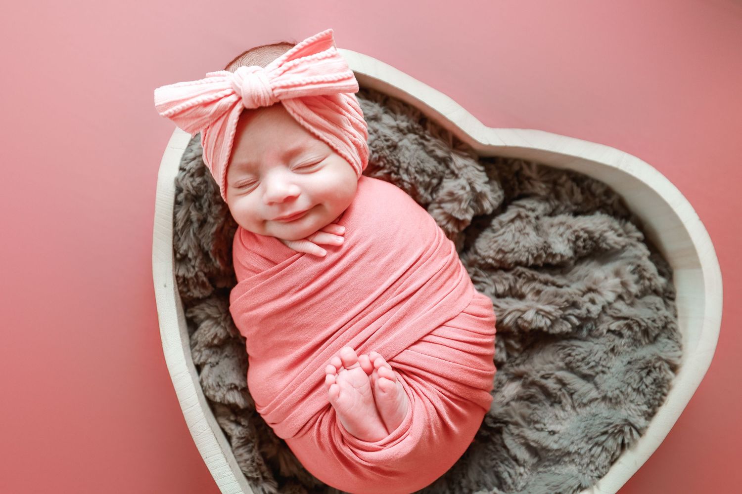 Smiling newborn baby swaddled in pink wrap with bow headband sleeping in heart-shaped bowl.