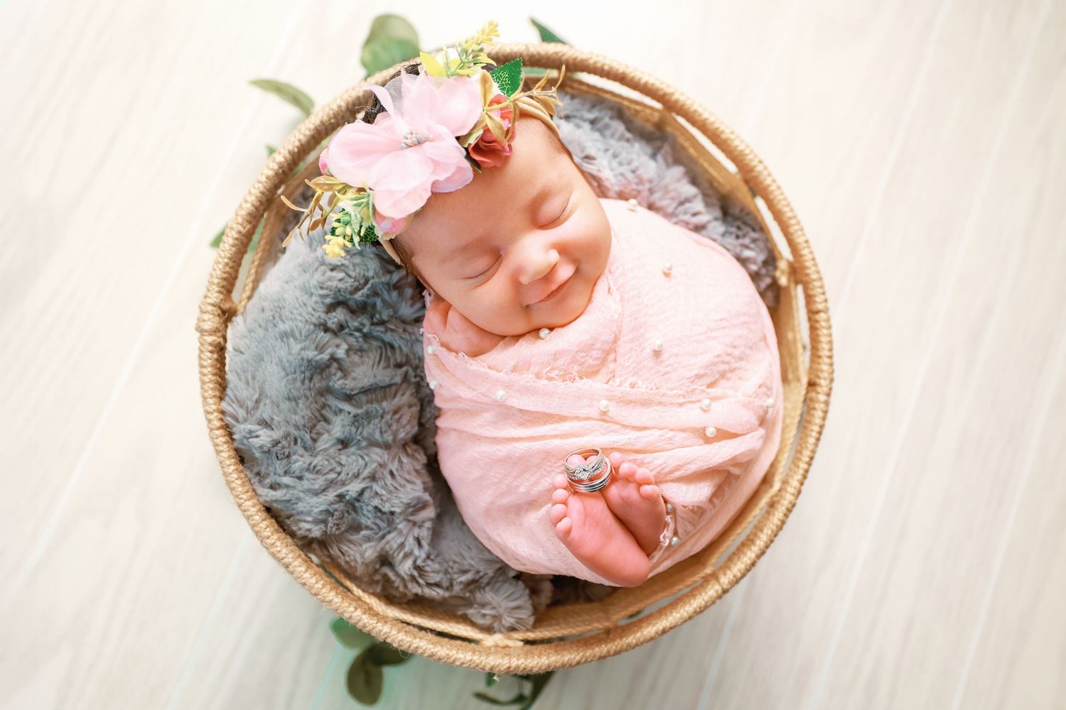 Smiling newborn baby wrapped in pink swaddle with floral headband sleeping in a wicker basket with gray fur.