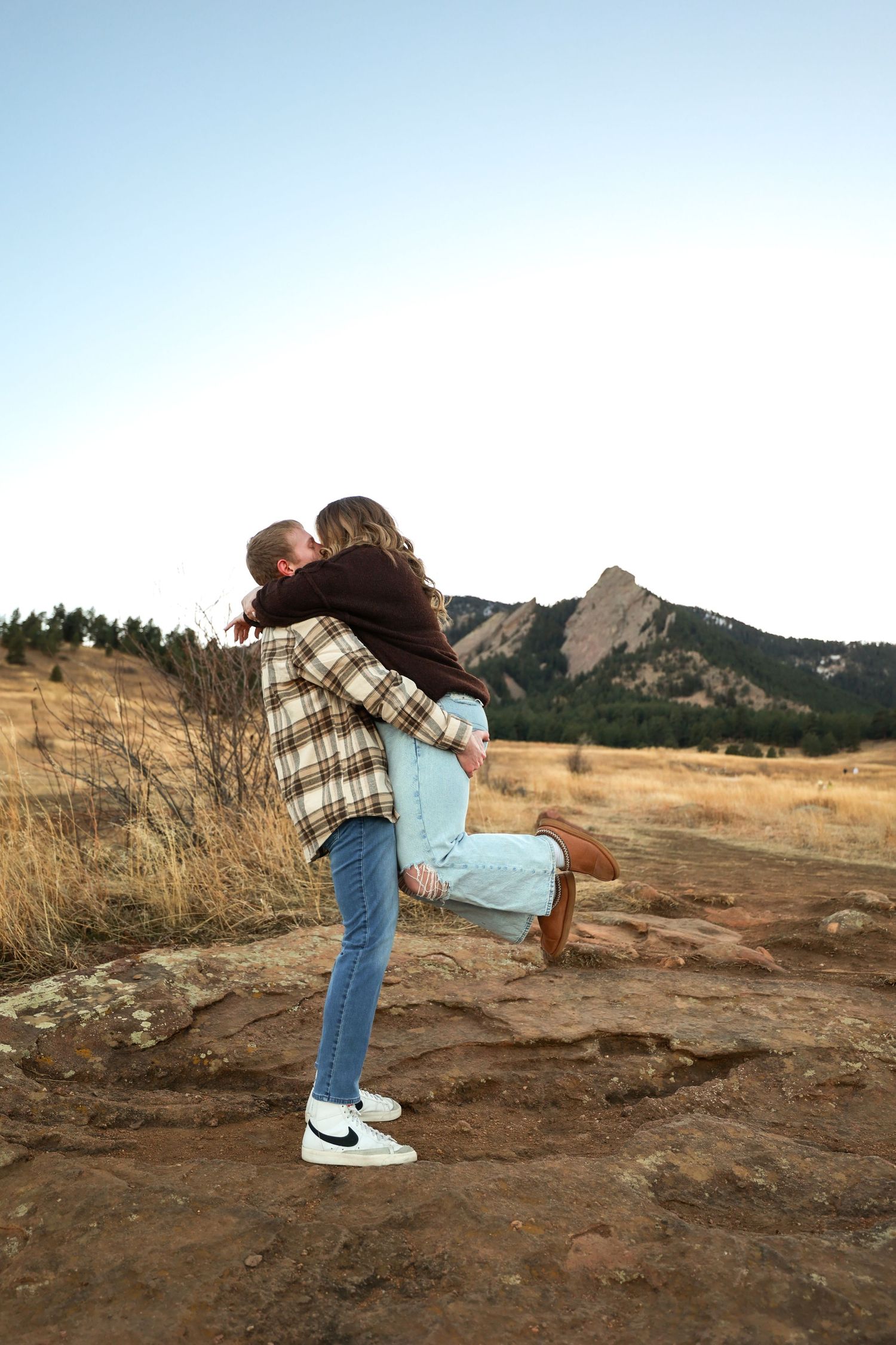 Man lifting and kissing woman near rocky terrain with Flatiron mountains in background.