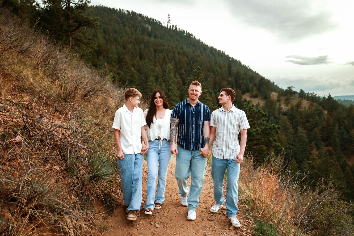 Family of four smiling and walking on a mountain trail surrounded by pine trees and scenic hills.