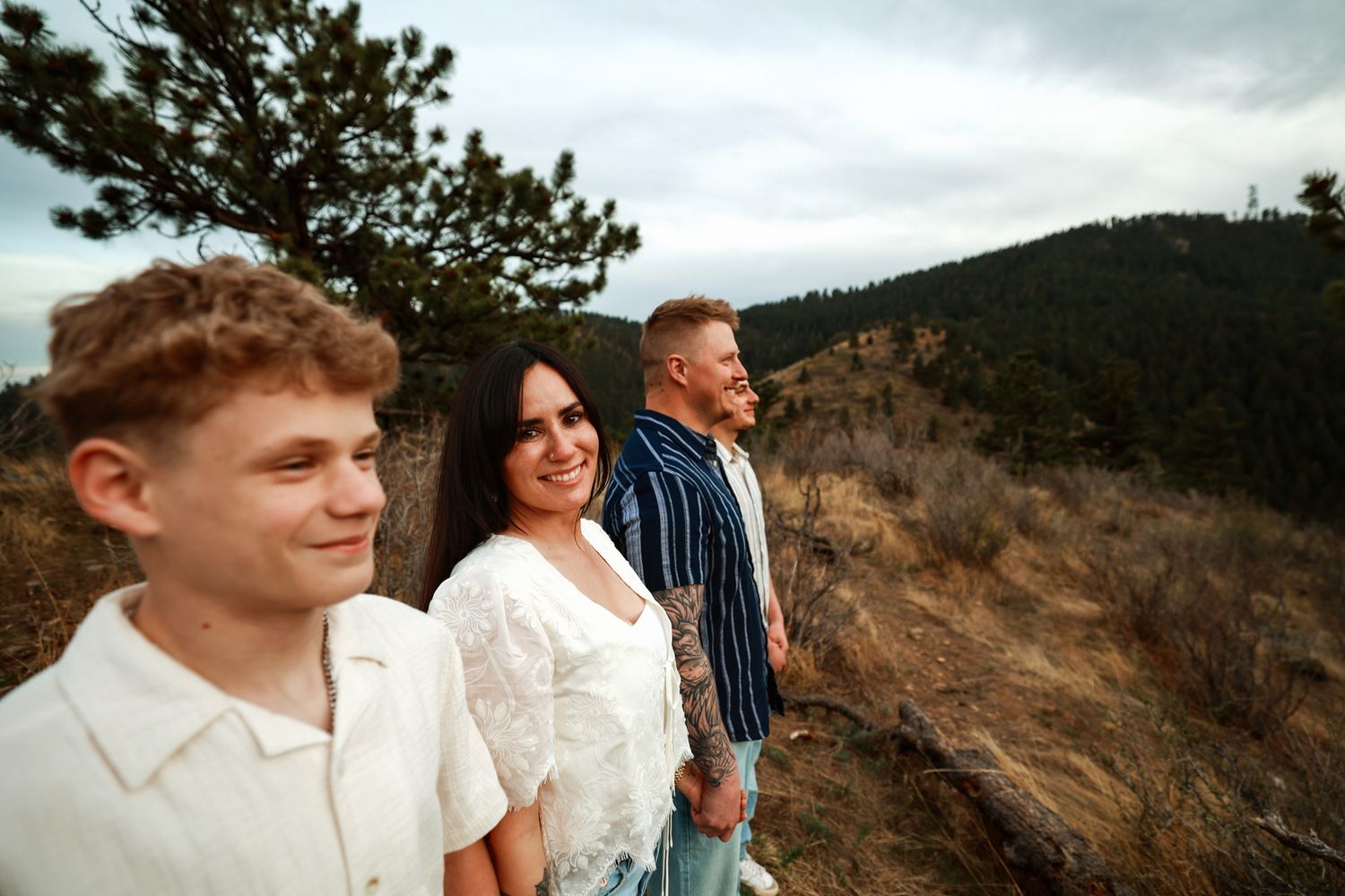 Family of three smiling on a scenic mountain overlook surrounded by pine trees and rolling hills.