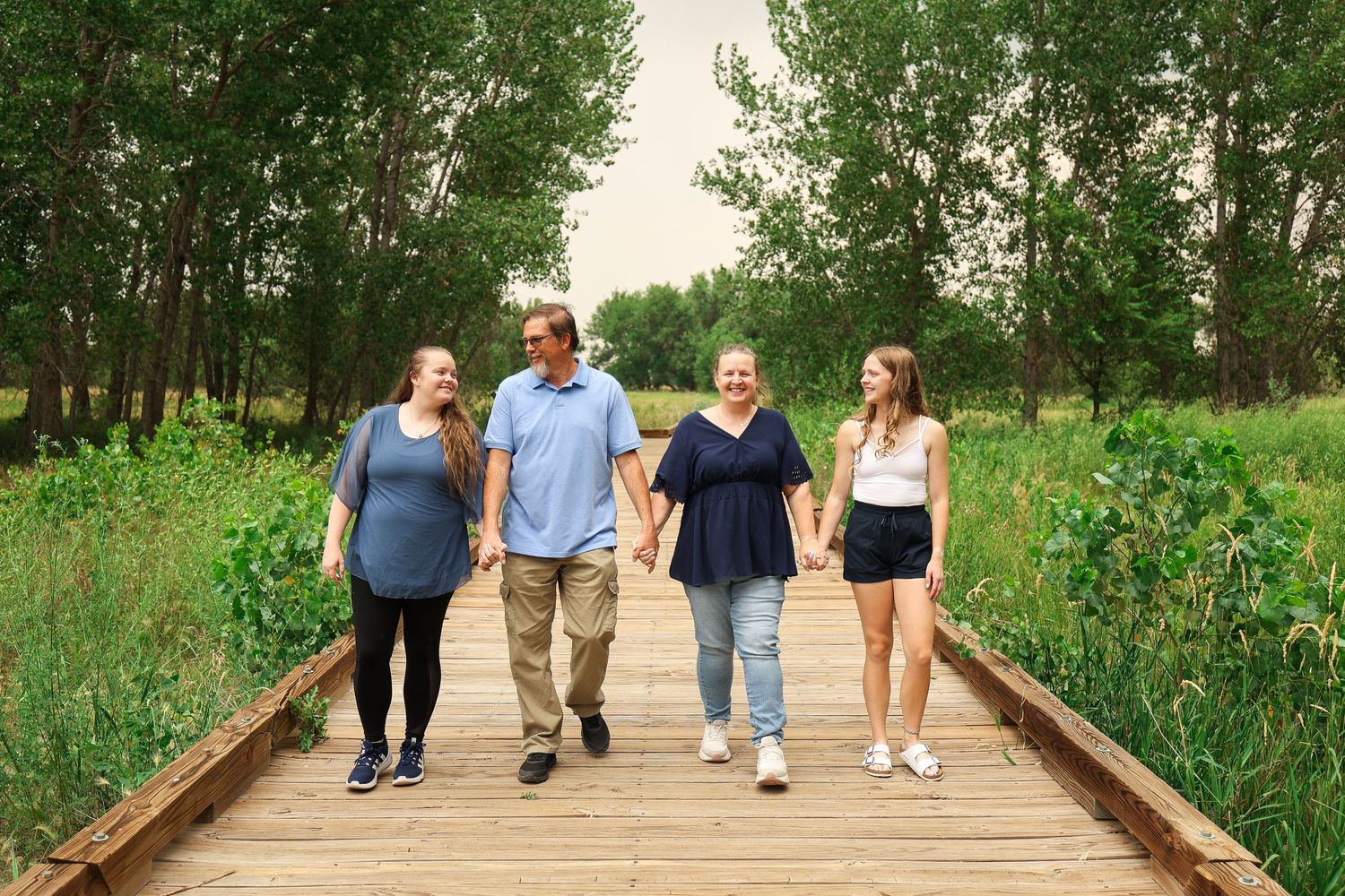 Family of four walking hand in hand on wooden boardwalk path through lush green summer forest.