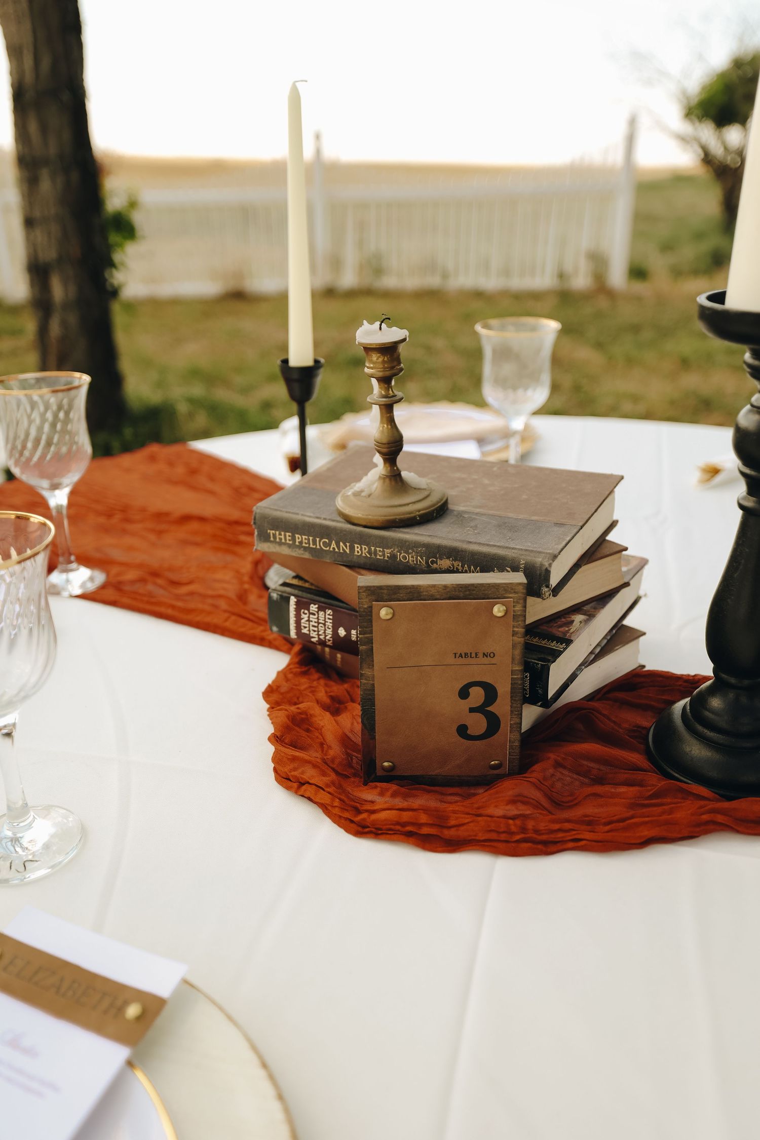 Elegant wedding table centerpiece featuring stacked vintage books, table number 3, and tall candlesticks on a rust runner.