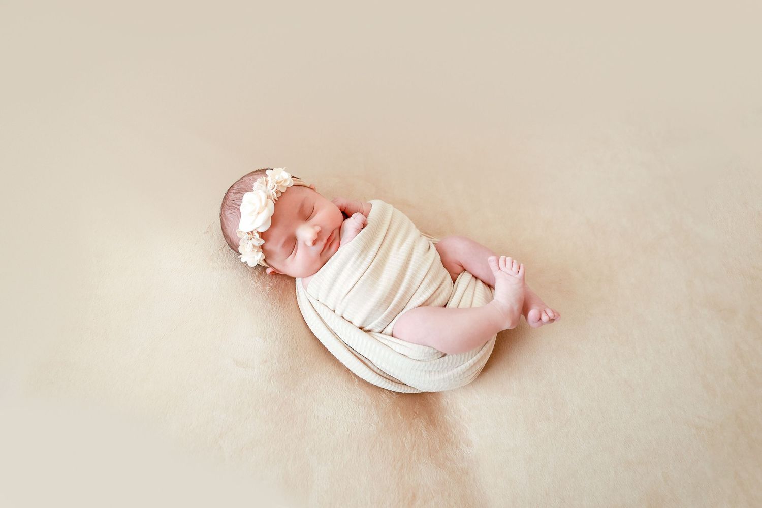 Newborn baby girl wrapped in cream swaddle with floral headband lying on soft beige fur blanket.