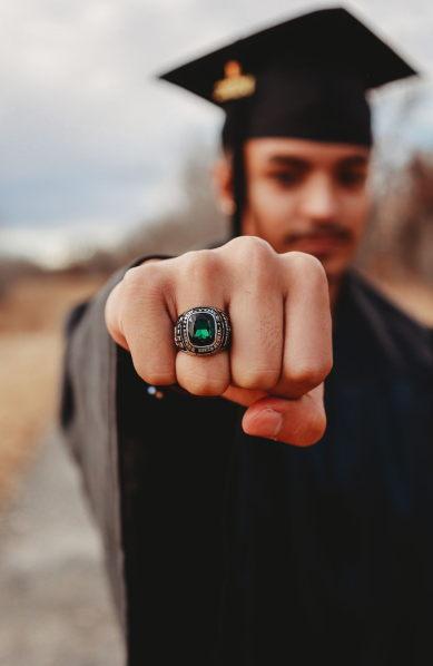 Male graduate in cap displays a green gemstone class ring close to the camera outdoors.