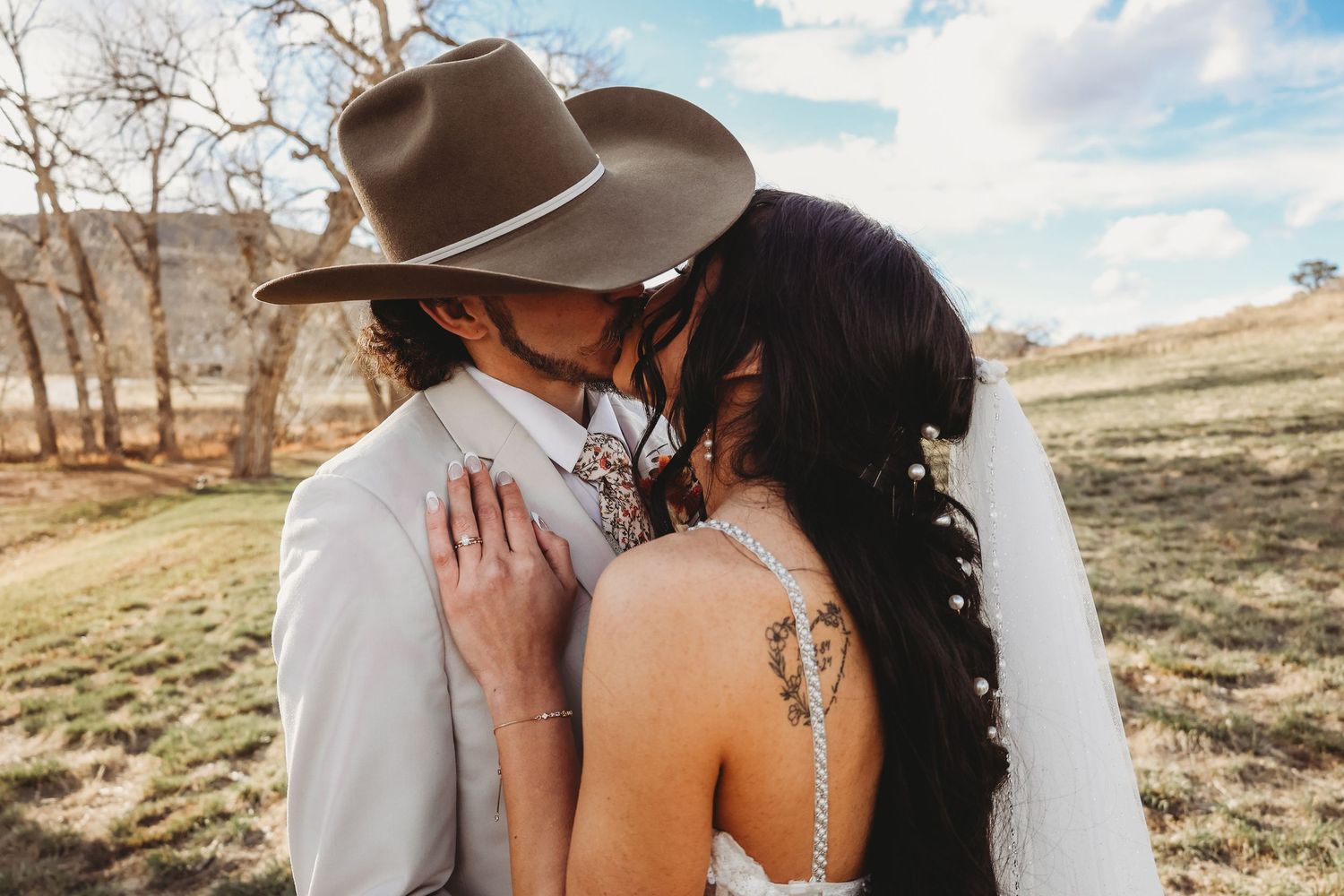 Bride and groom sharing a kiss outdoors, groom wearing a cowboy hat and white suit, bride in a backless wedding dress with tattoos.