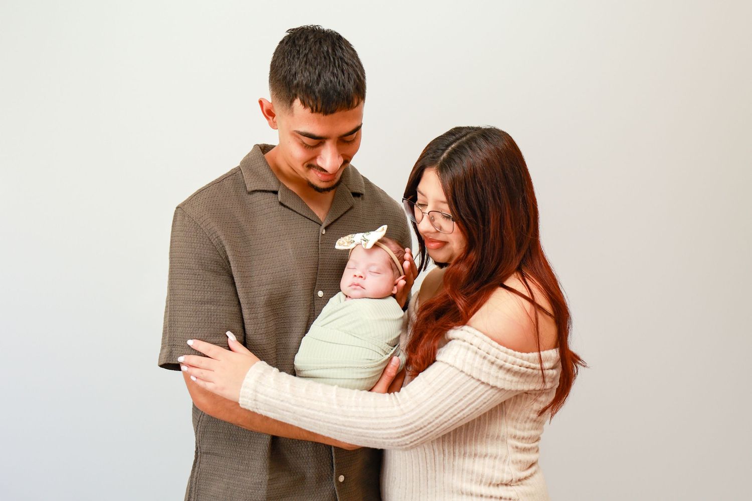 Young couple smiling at their newborn baby dressed in green, posed against a white background.