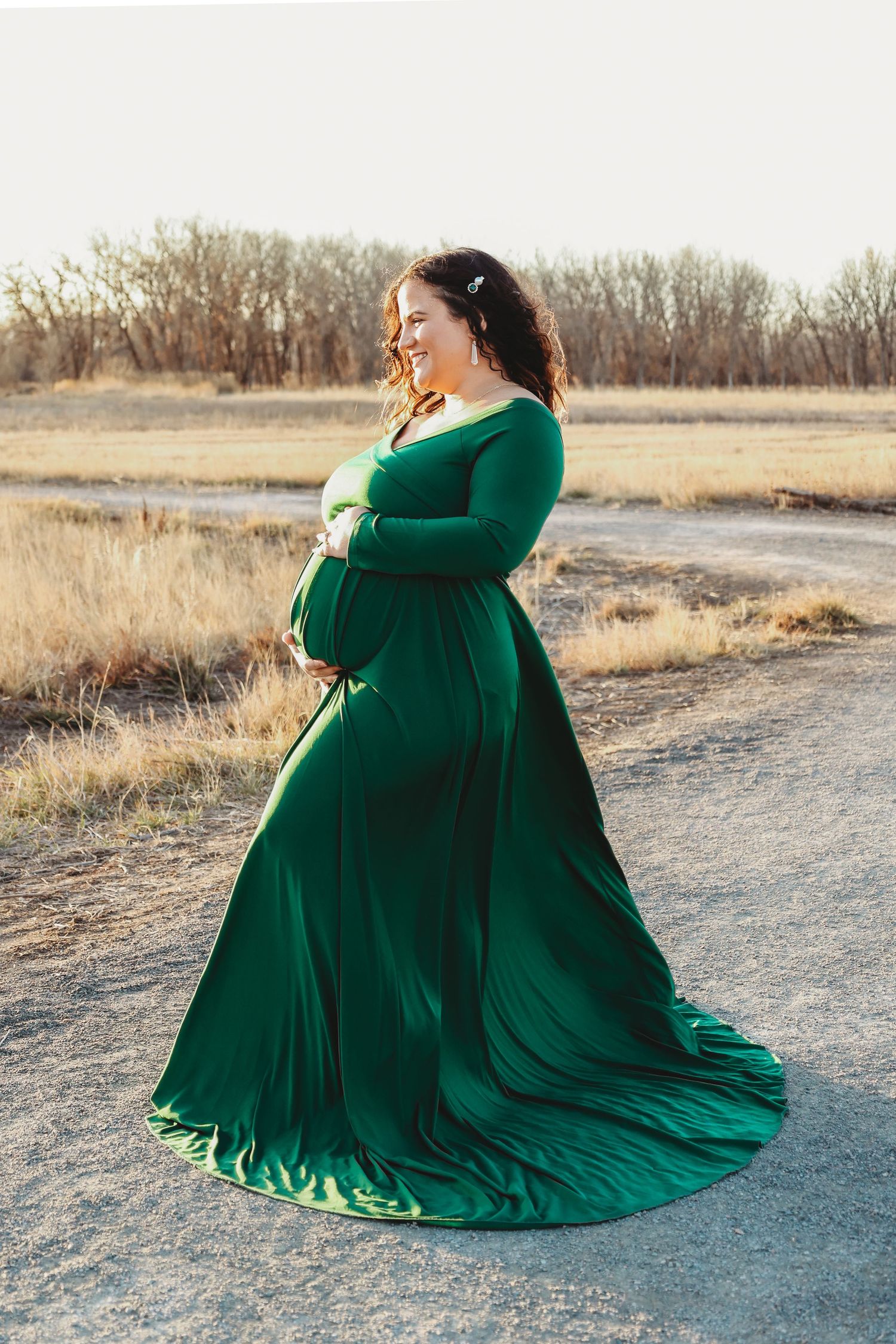 Pregnant woman in elegant green maternity gown standing outdoors in a field at golden hour.