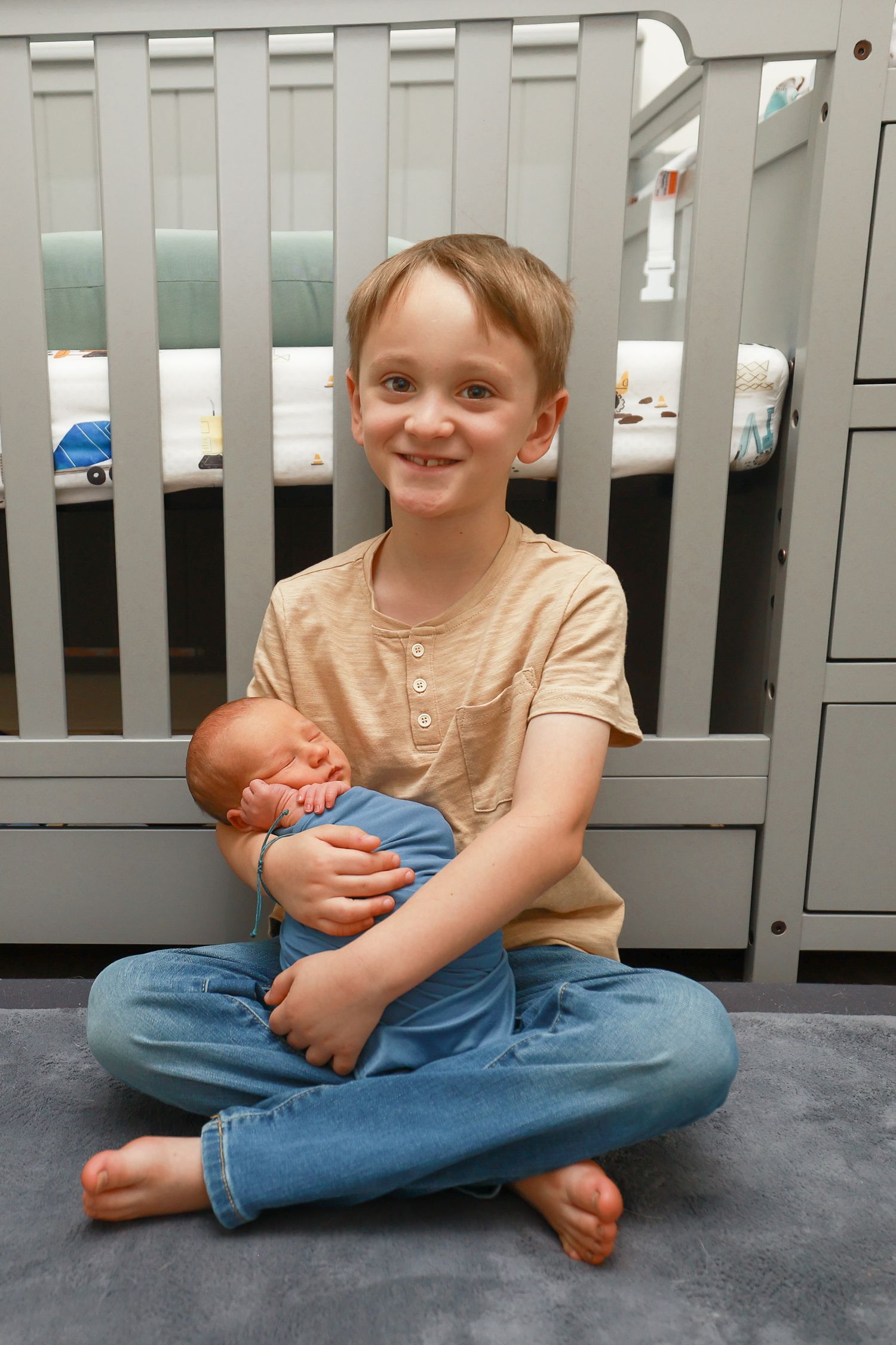Smiling older brother proudly holds his swaddled newborn sibling while sitting cross-legged in front of a crib.