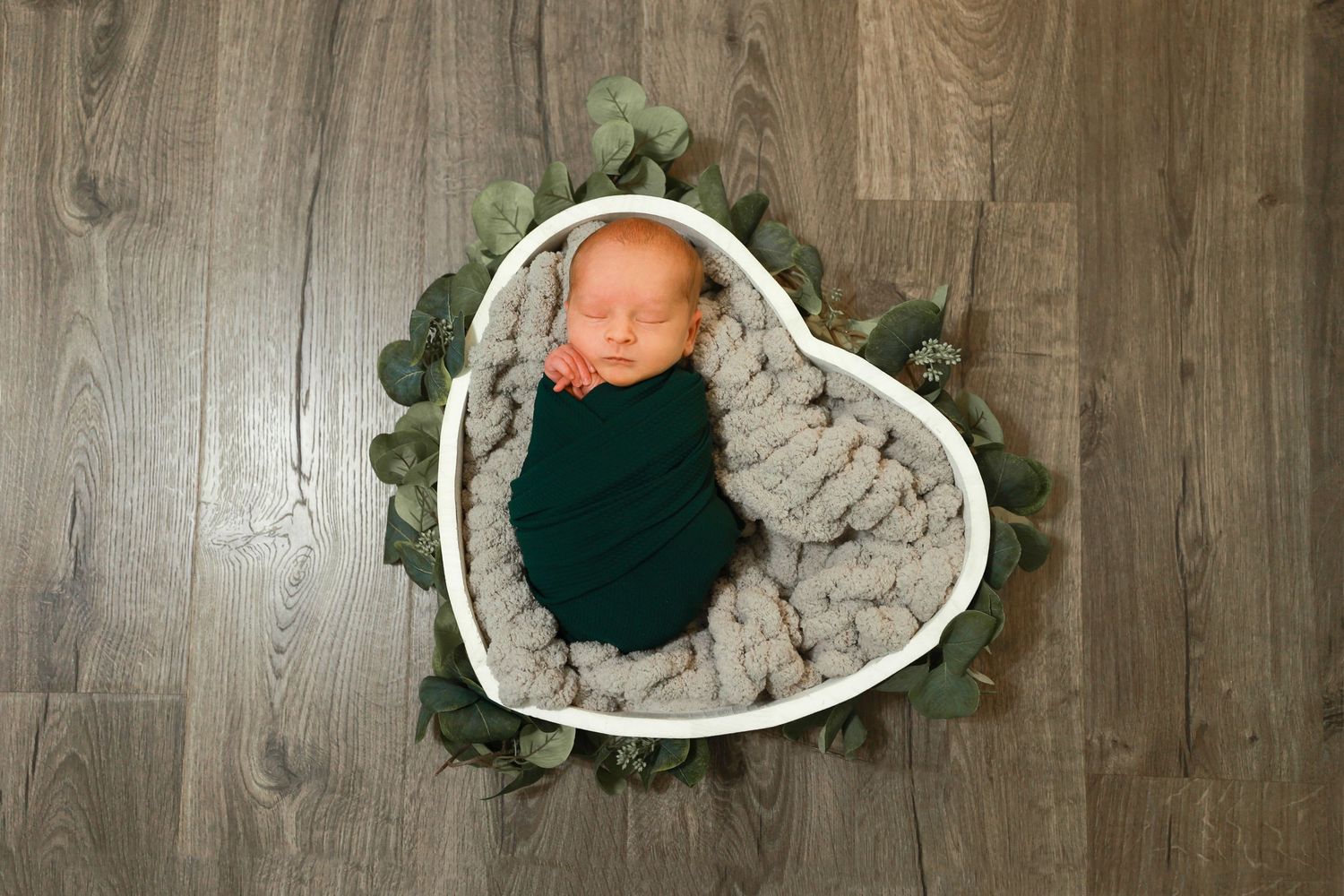 Peaceful newborn baby wrapped in dark green swaddle resting in a heart-shaped bowl.