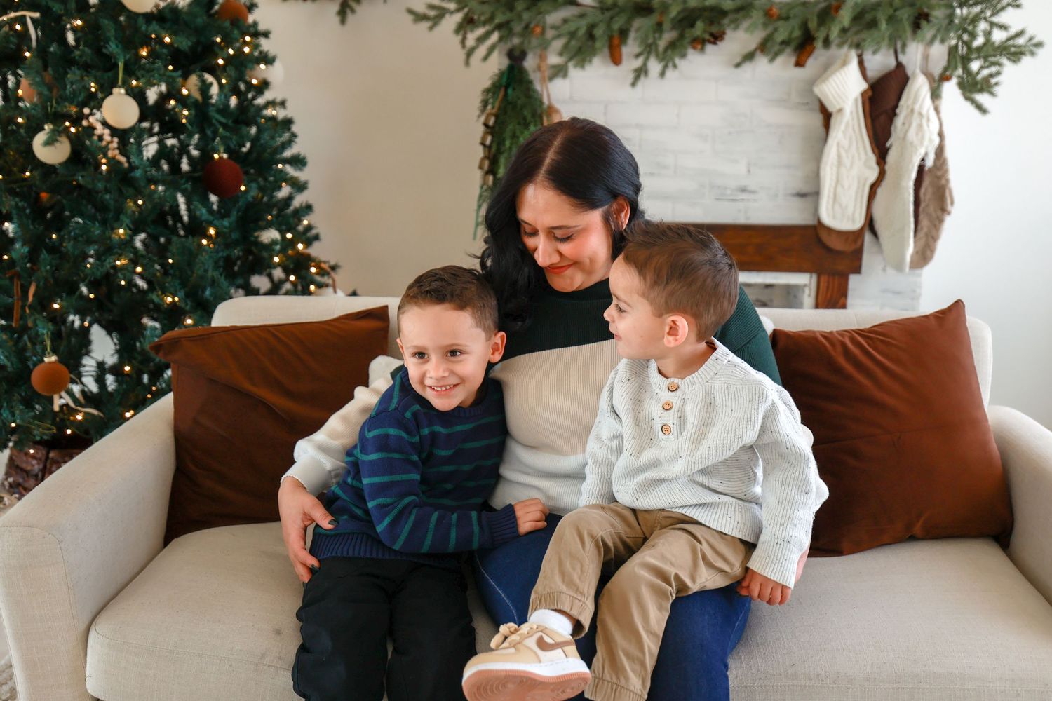 Mother sits on cream sofa hugging two young boys with Christmas tree and holiday decorations in background.