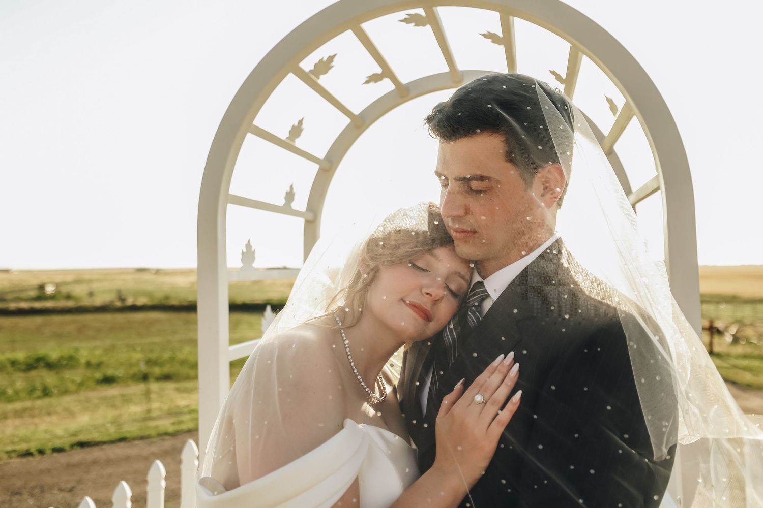 Romantic bride and groom embrace under white arched arbor at golden hour farm wedding.