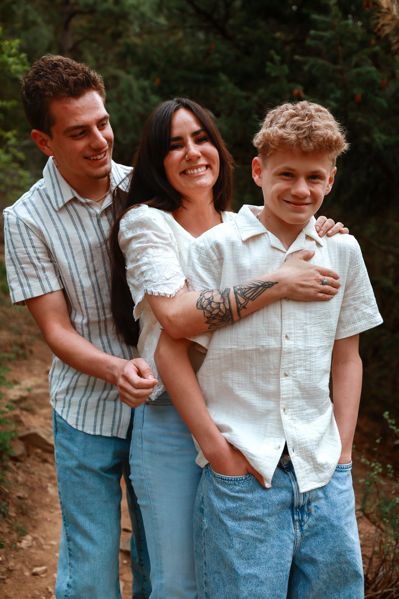 A happy family of three poses outdoors, smiling together among green trees in casual blue jeans and white shirts.