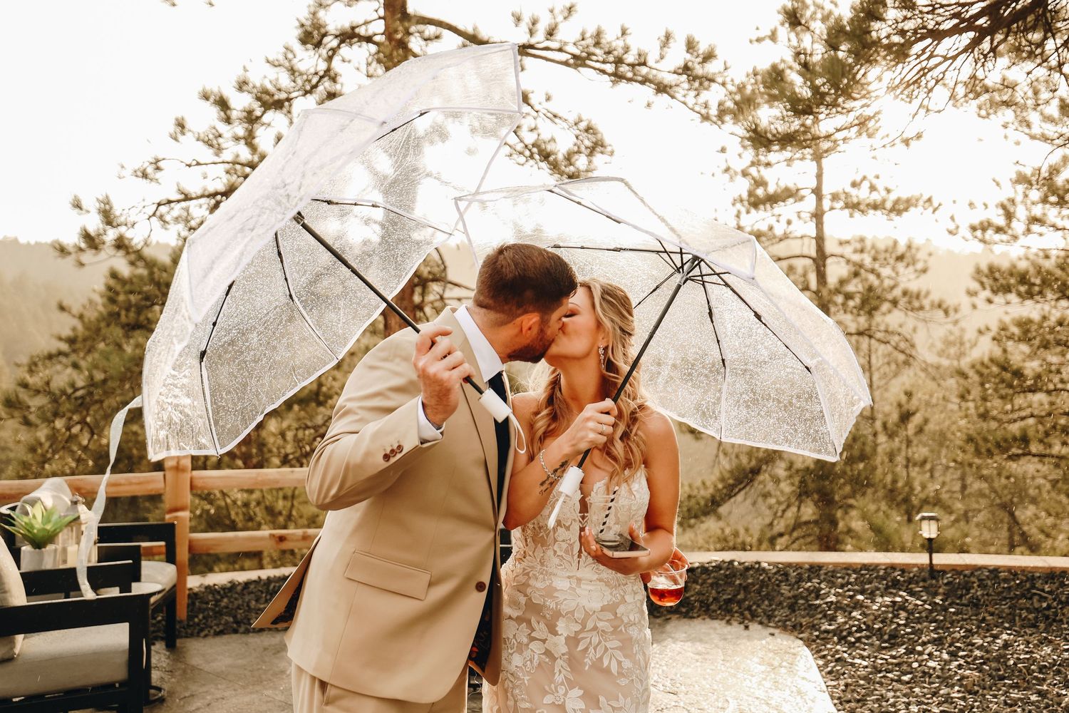 Bride and groom kissing under clear umbrellas in outdoor wedding ceremony surrounded by pine trees.