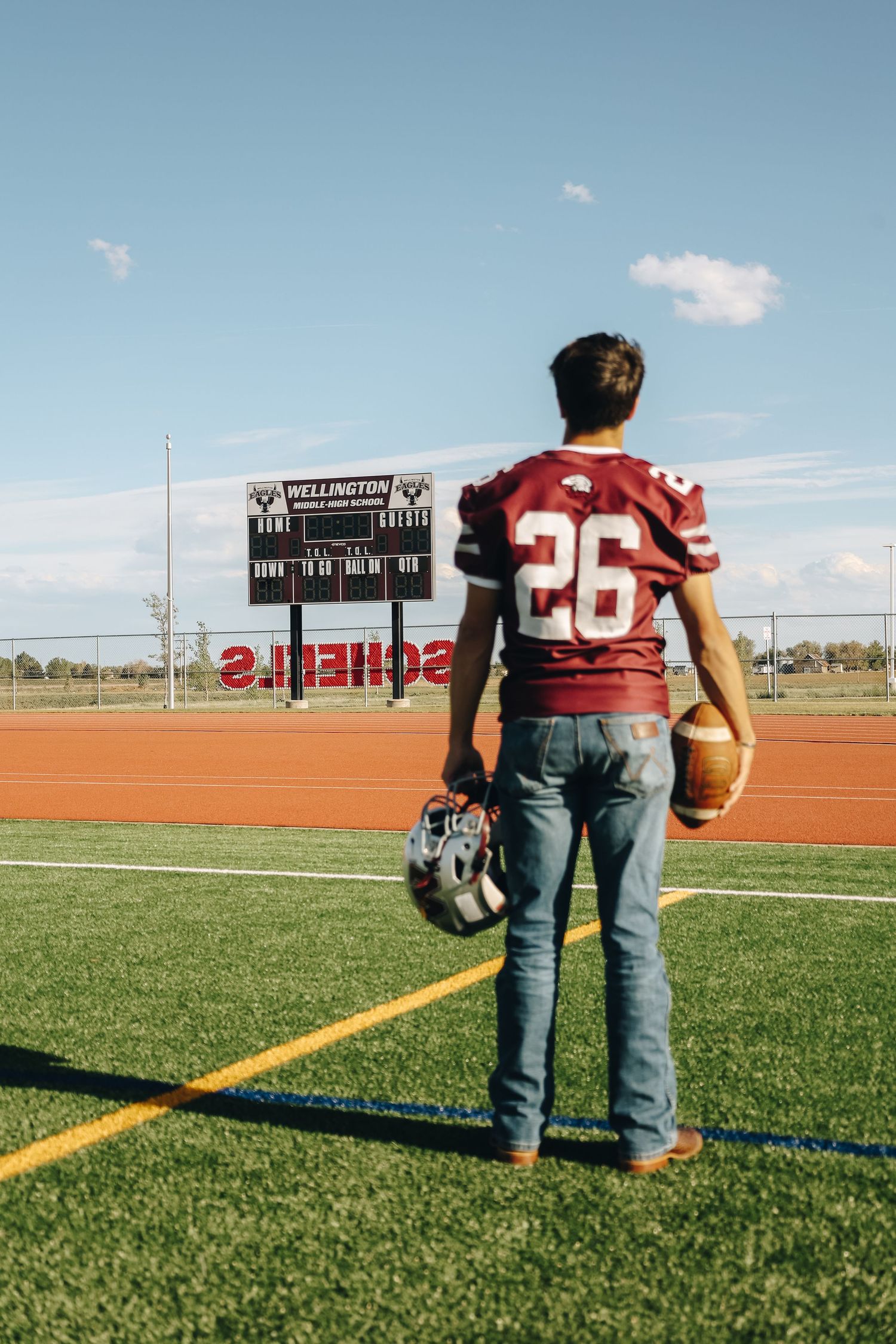Football player in maroon number 26 jersey holding helmet facing scoreboard on athletic field.