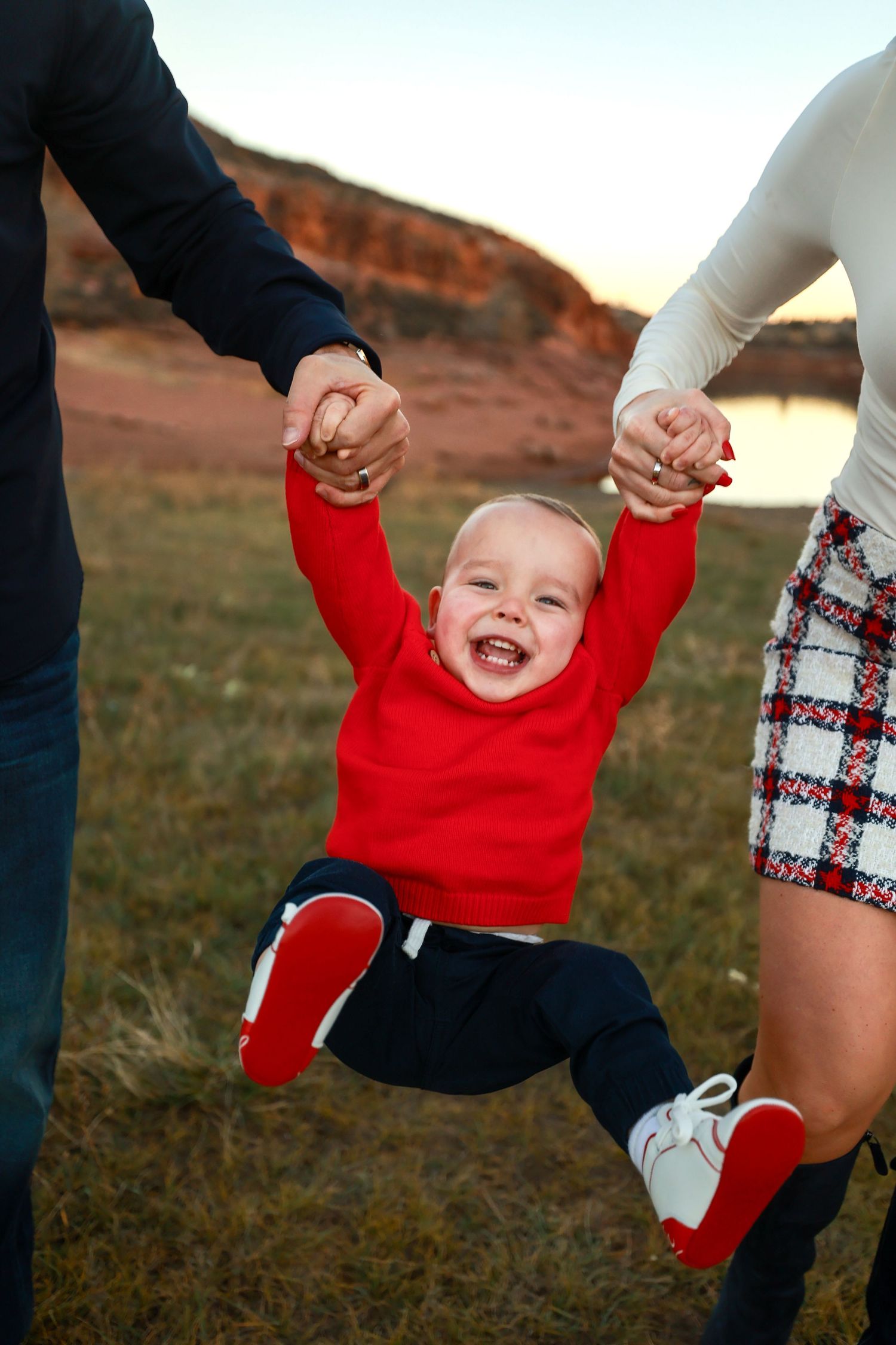 Laughing toddler in red sweater being swung by parents hands near scenic red rock landscape.