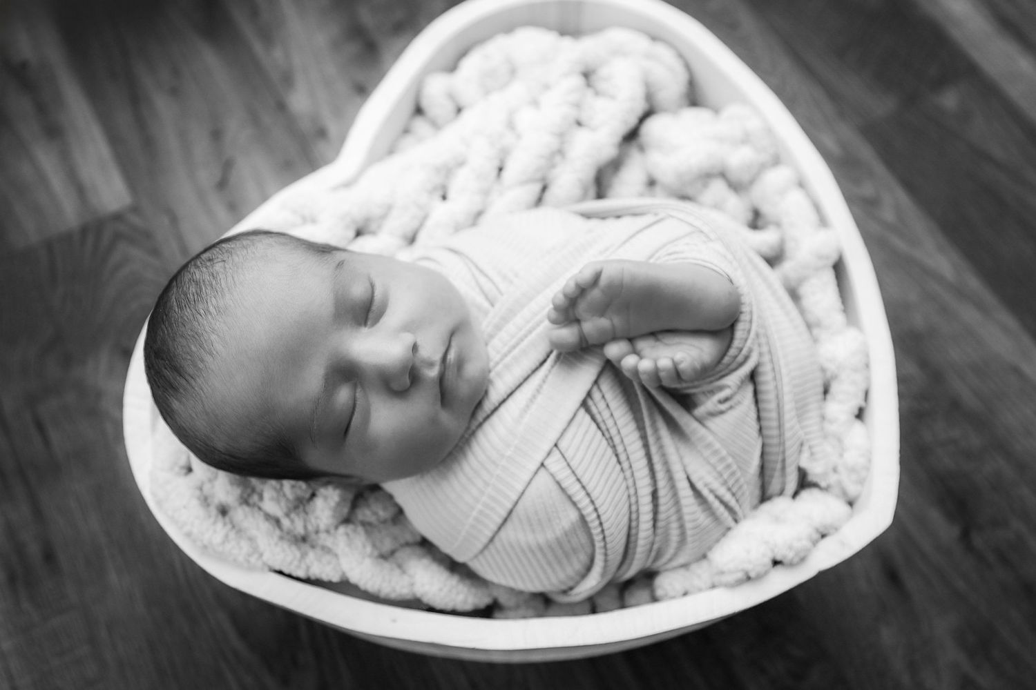 Black and white photo of a sleeping newborn baby swaddled in a heart-shaped bowl with a chunky knit blanket.