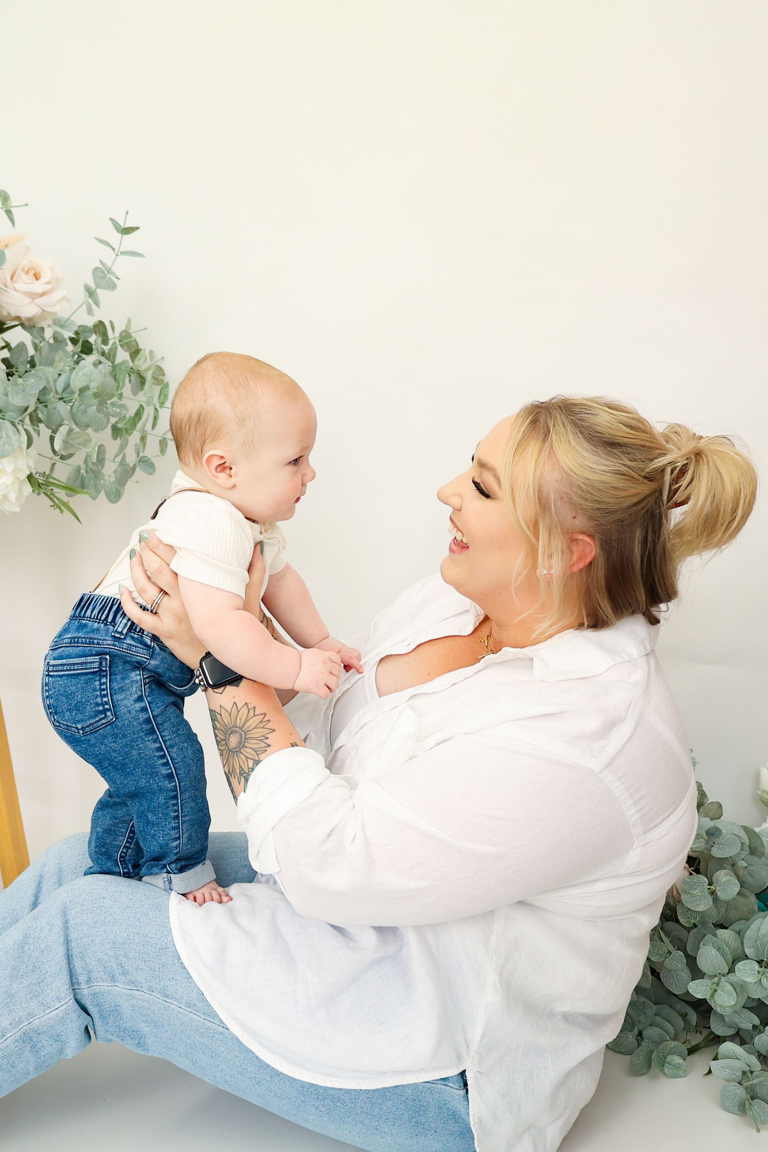 Smiling blonde mother in white shirt holding her baby upright during a clean white studio photoshoot.