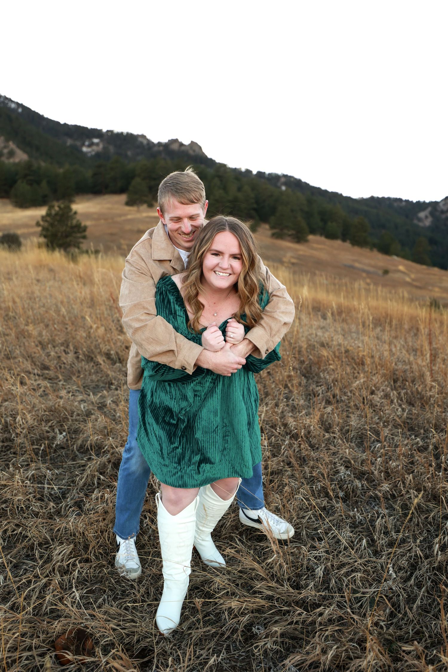 Happy couple hugging in dry grass field with mountain backdrop, woman wearing green velvet dress and white boots.