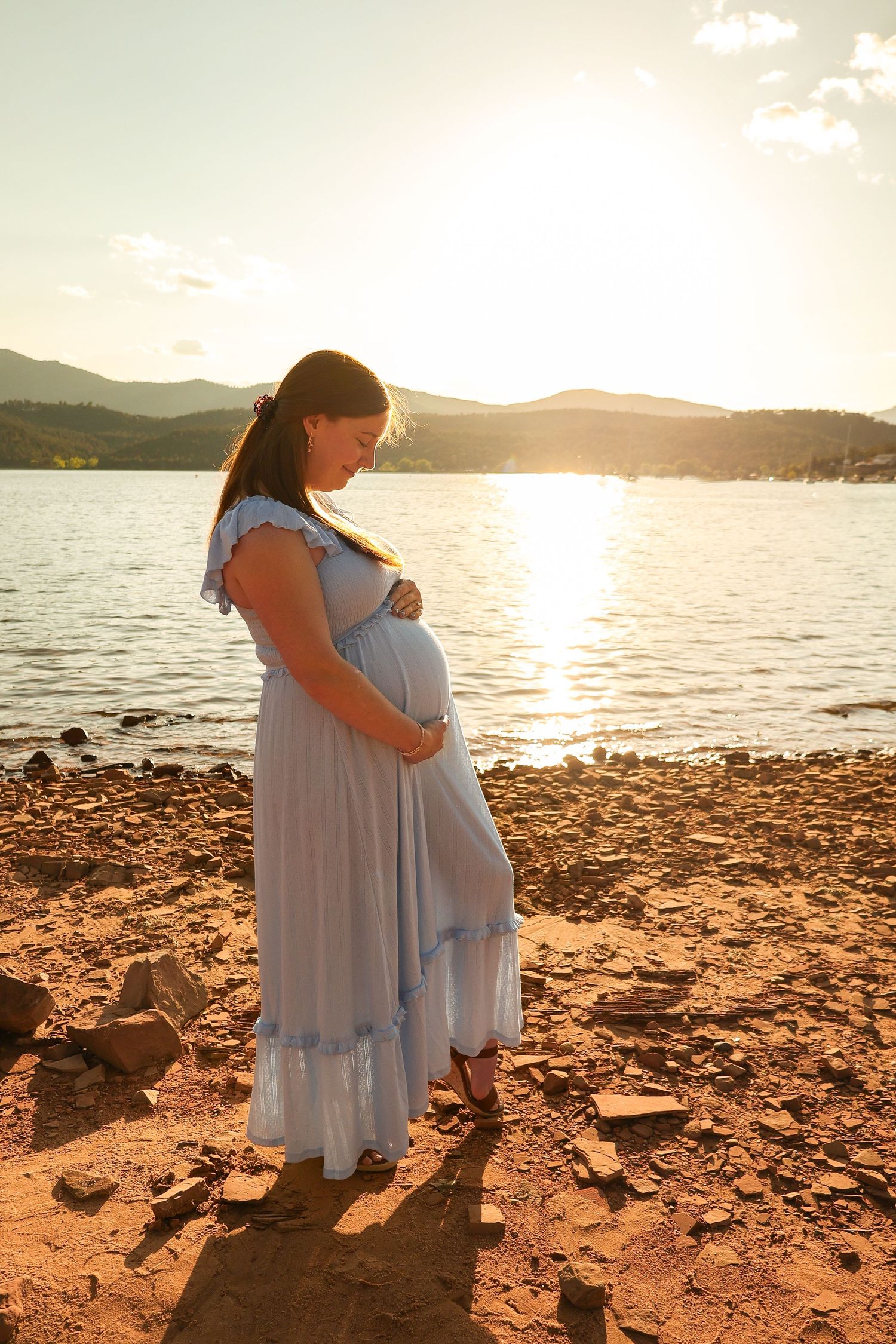 Pregnant woman in flowing blue dress cradles baby bump during golden sunset maternity session by lakeside.
