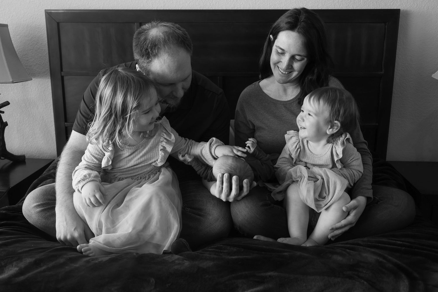 Black and white family photo of parents and two young children sitting on bed.