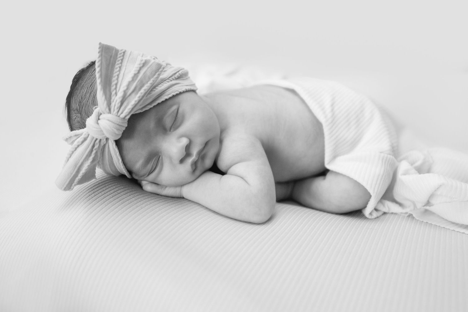 Black and white portrait of sleeping newborn baby girl with bow headband resting on hands on soft blanket.