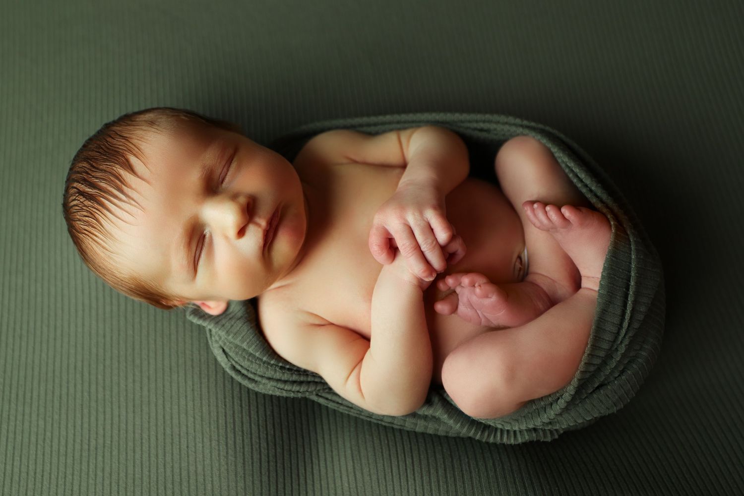 Sleeping newborn baby boy unwrapped from a green knit wrap on a matching green backdrop.
