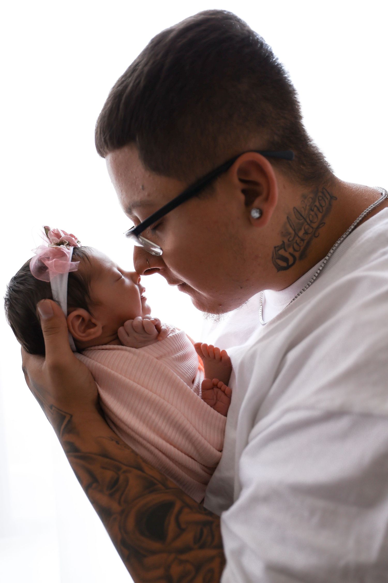 Tattooed parent tenderly holding a newborn baby girl in a pink swaddle nose-to-nose.