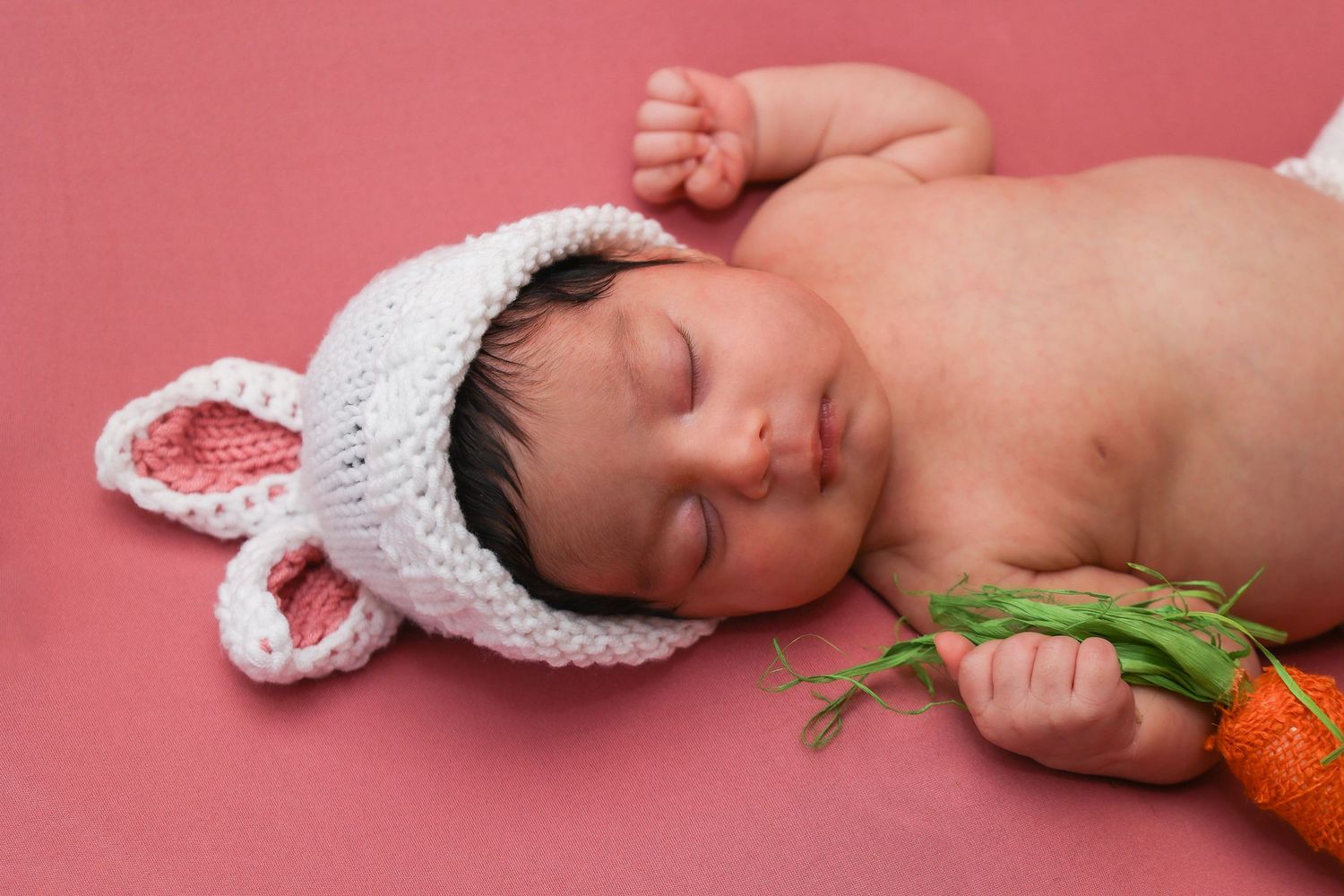 Sleeping newborn baby wearing knit bunny hat holding a carrot on pink background.