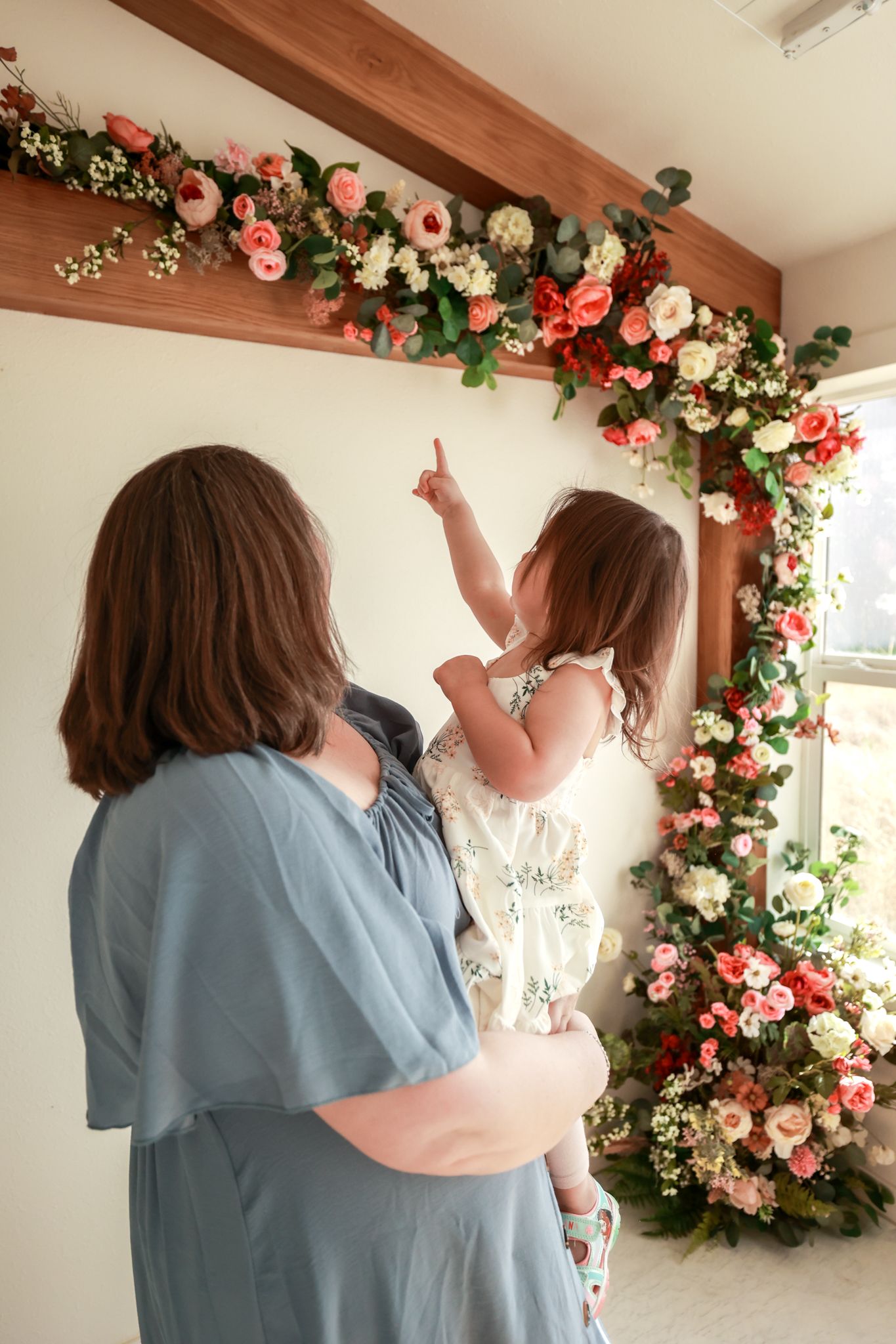 Woman in blue dress holds young girl pointing at floral arch with pink and white roses indoors.