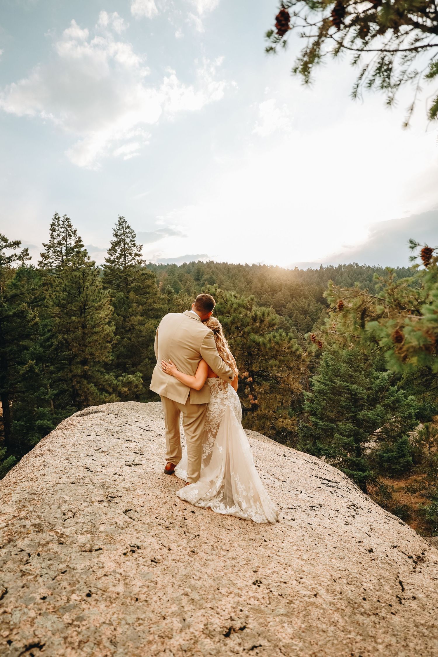 Bride and groom embracing on a rocky cliff surrounded by pine trees at sunset in a romantic outdoor wedding photo.