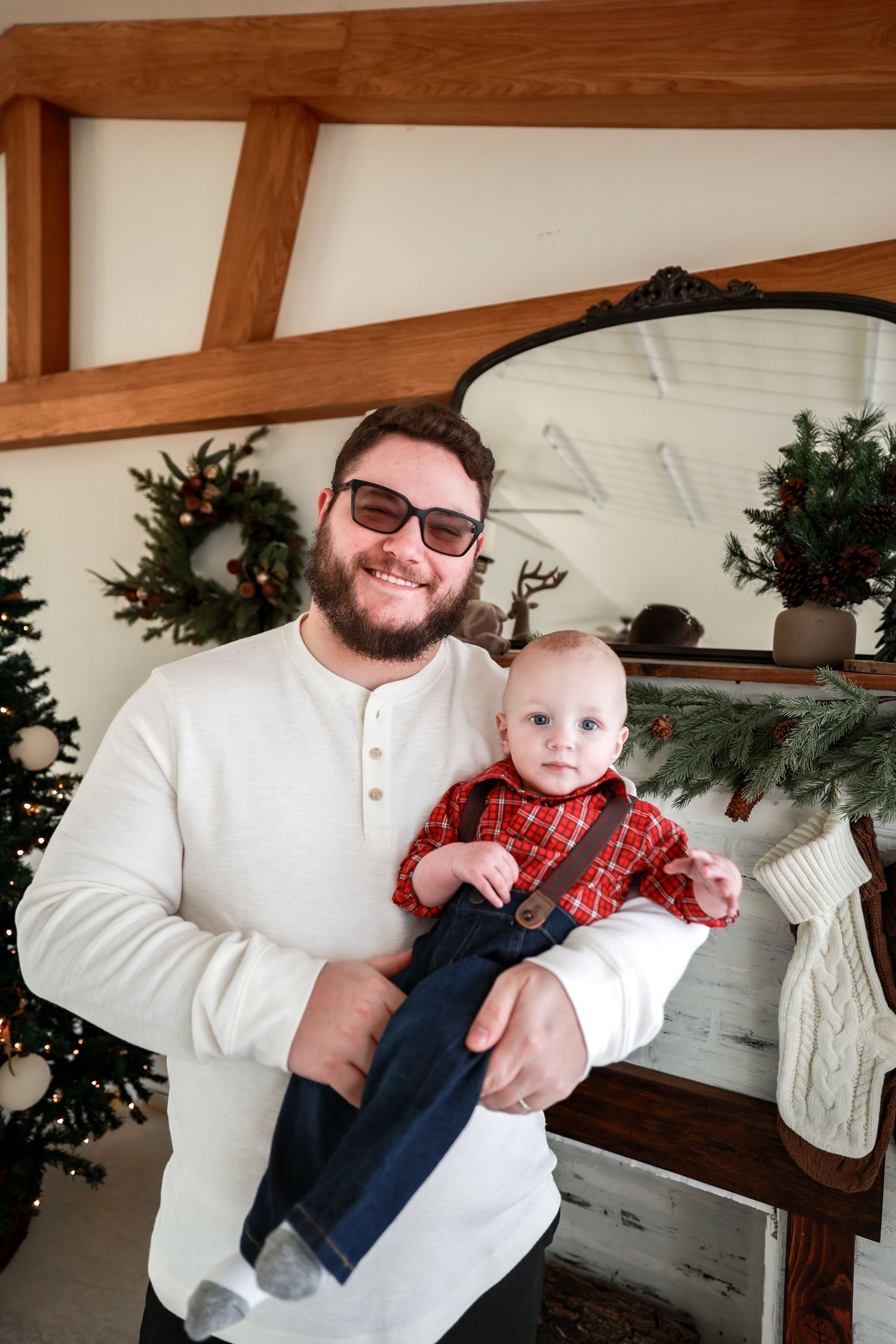 Bearded man in white sweater and sunglasses holds baby dressed in red plaid outfit near Christmas tree.