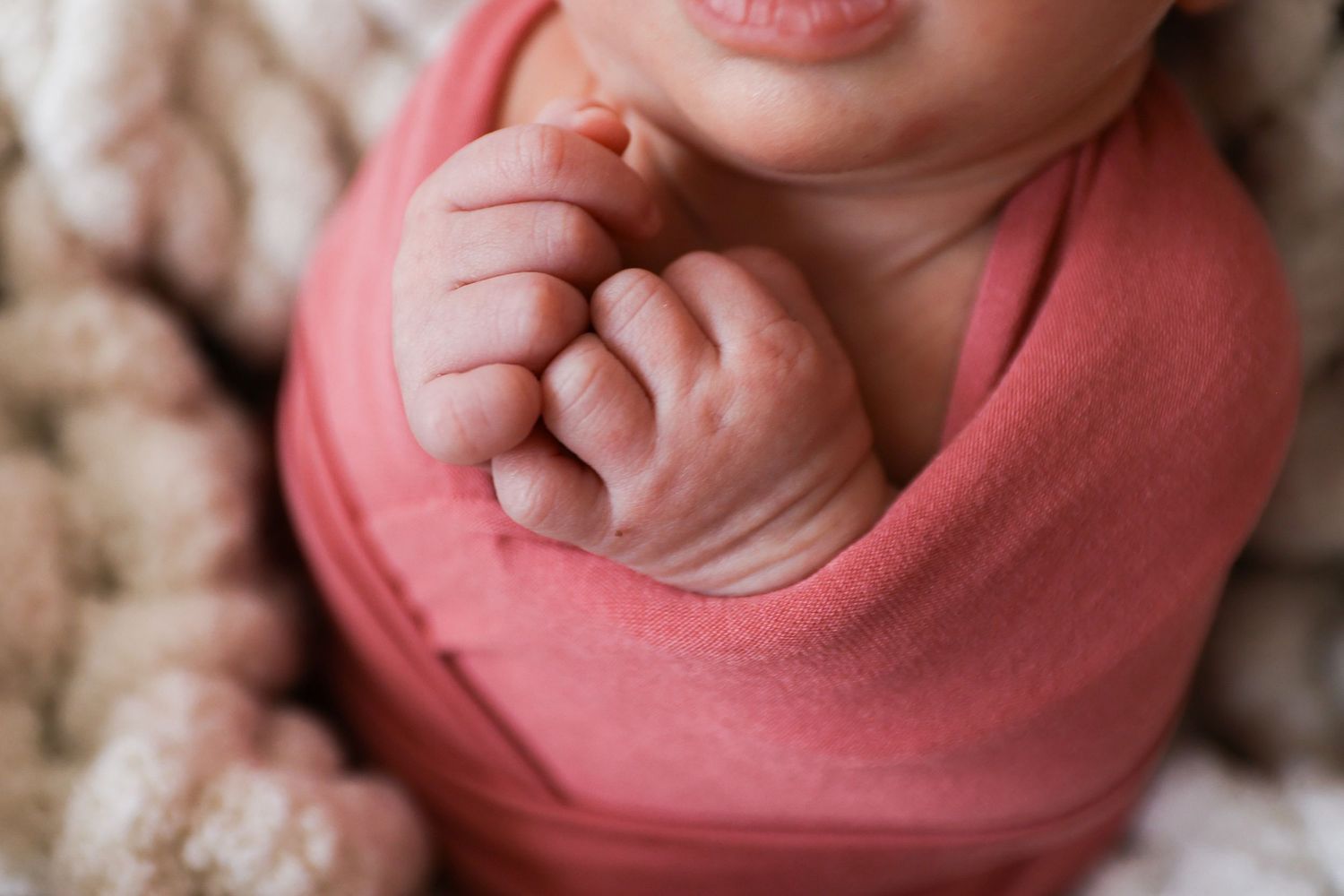 Close-up of a swaddled newborn baby in a coral pink wrap with tiny hands visible.