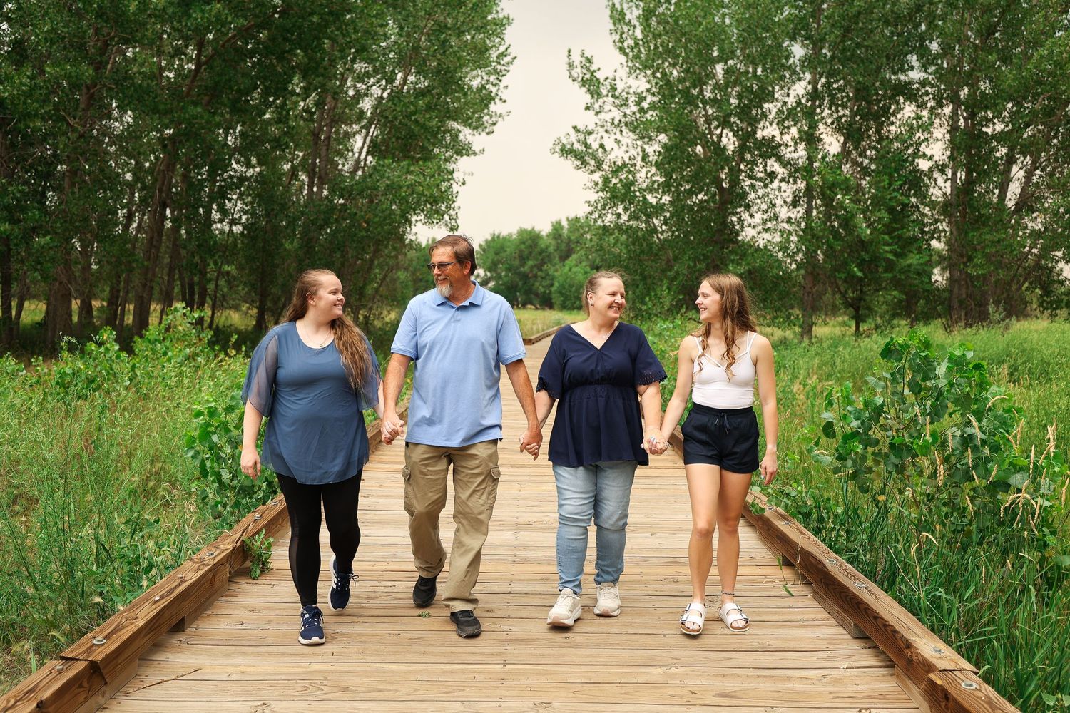 Family of four walking hand in hand on wooden boardwalk path through lush green nature trail.