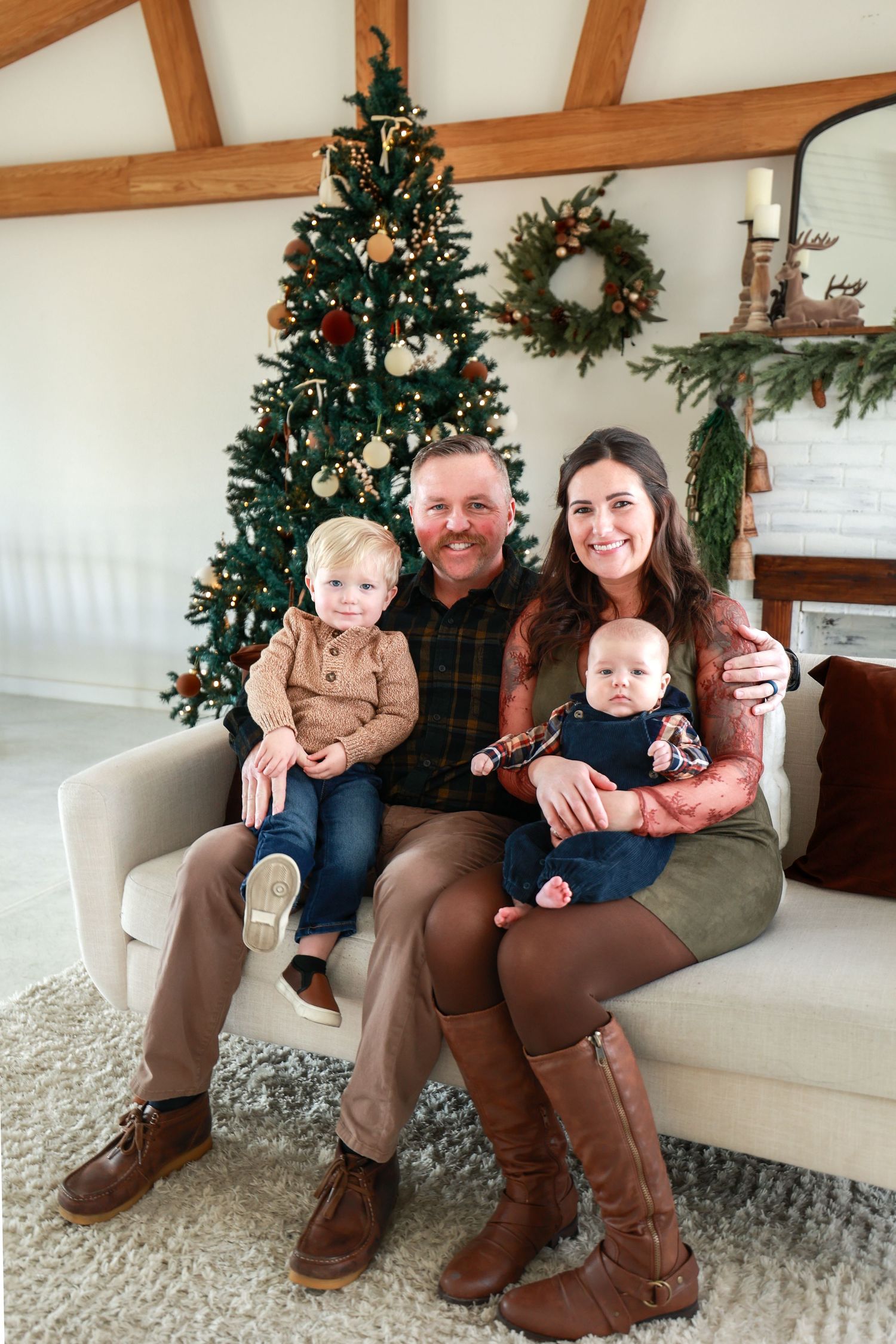 Family of four with toddler and baby sit on sofa in front of decorated Christmas tree in bright living room.