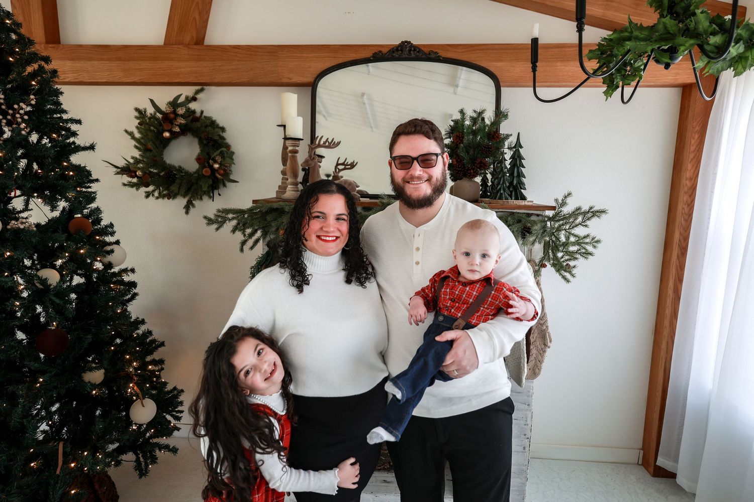 Family of four with two children smile together in a decorated Christmas living room with wreaths and tree.