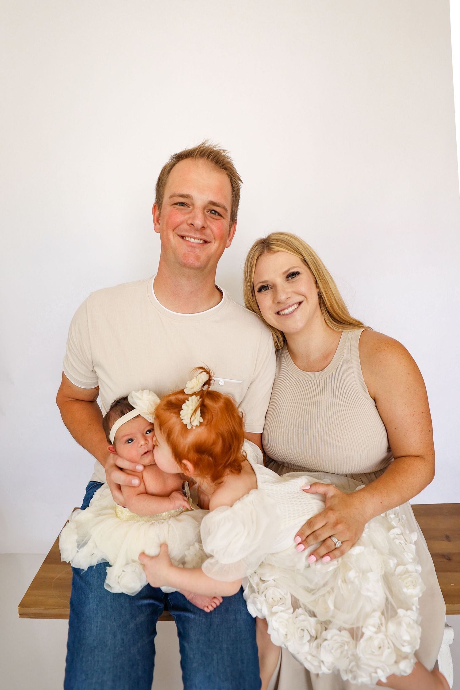 Smiling parents sitting with red-haired toddler and newborn baby girl in white headband for family photo.